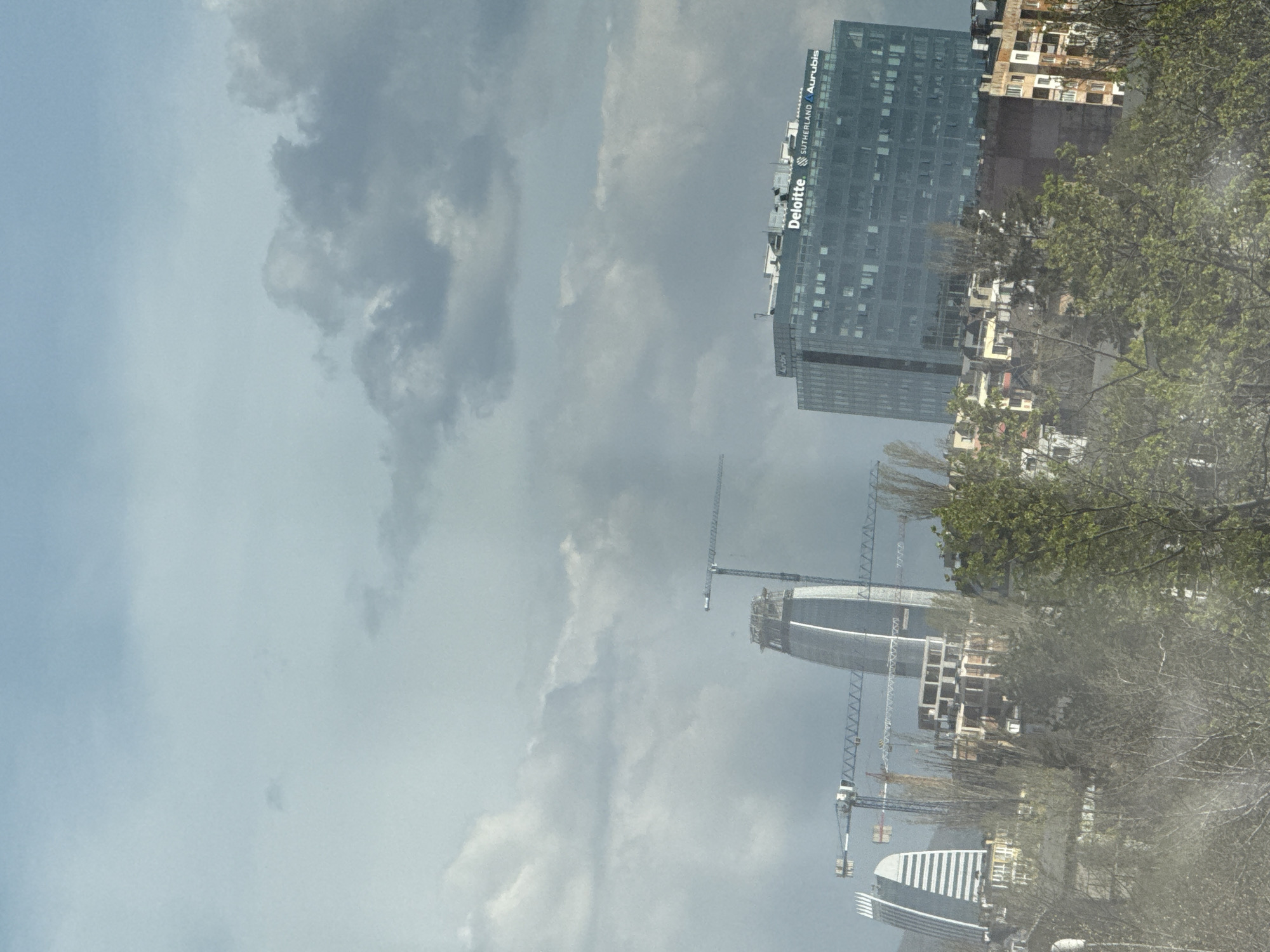 The image shows a view in Sofia, Bulgaria, featuring a cloudy sky above buildings and construction sites. Visible in the upper right is a tall office building with "Deloitte Sutherland Aurubis" signage. Below and further to the left are two buildings under construction, each surrounded by cranes. The foreground includes trees with some leaves.