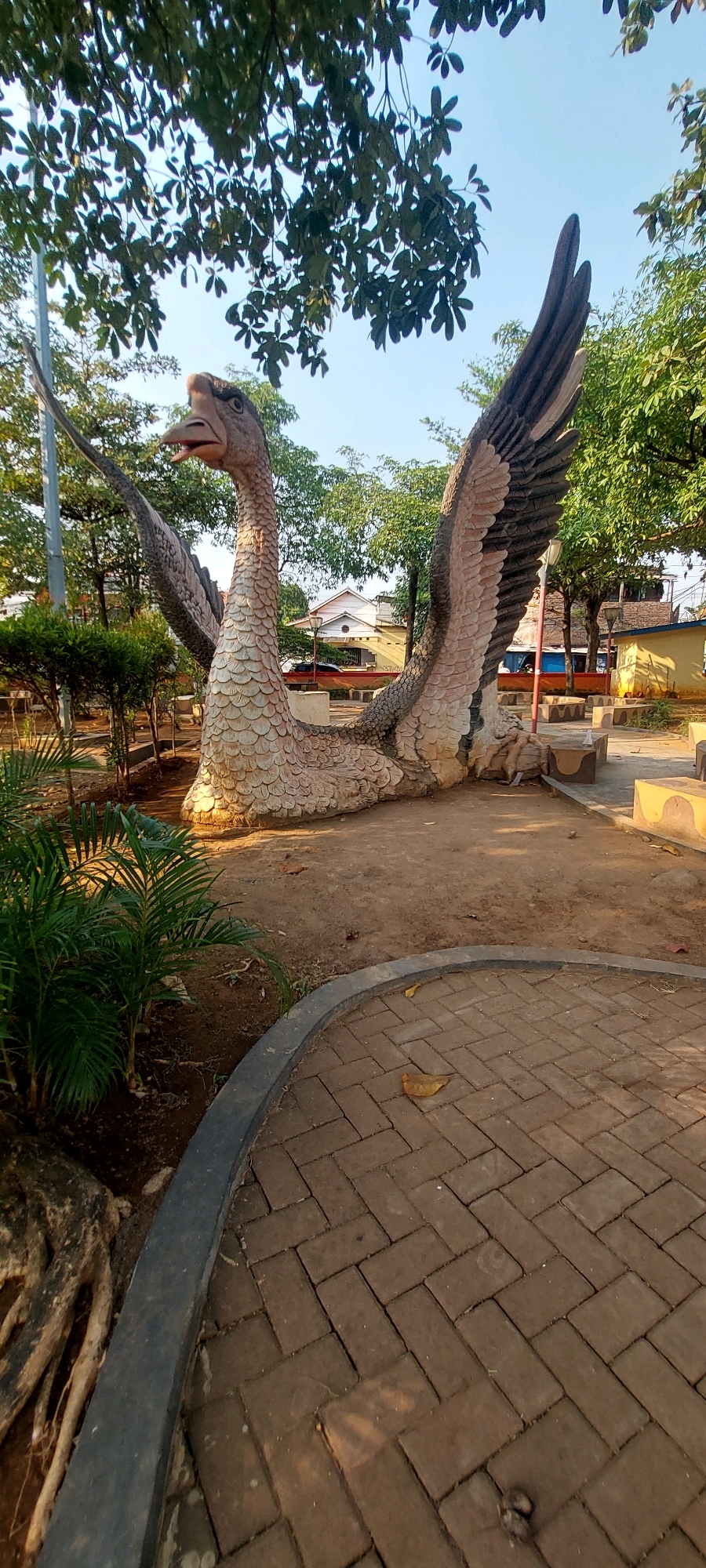 The image shows a large concrete statue of a prehistoric bird, possibly a prehistoric waterfowl or a theropod dinosaur with bird-like features, resting on the ground in a park. The statue is detailed, with scales on its body and feathers on its wings. It has a long neck and an open beak, giving it a somewhat fierce or surprised expression. The statue is located in an outdoor public space, with paved walkways made of brick, benches, and surrounding greenery including trees and smaller plants. In the background, there are buildings, indicating an urban or suburban setting. The sky is clear and blue, suggesting it is daytime. The overall impression is of a park designed with a theme, possibly related to ancient life or natural history. This location is likely the Taman Mpu Tantular in Semarang, Indonesia, a park that features various sculptures and historical replicas.