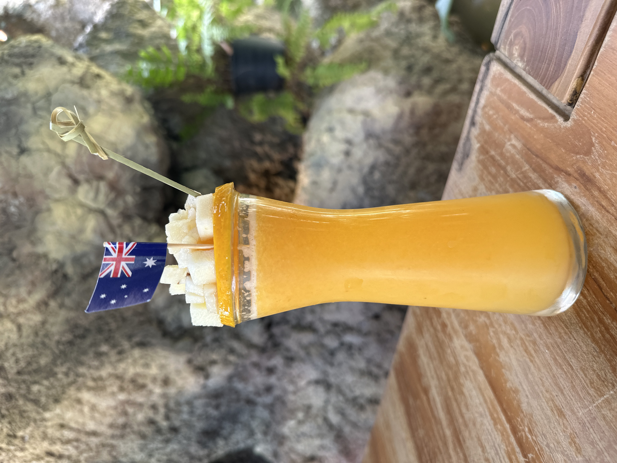 A close-up, eye-level shot features an orange smoothie in a clear glass, topped with small white cubes and a miniature Australian flag. The glass is placed on a wooden table with a visible grain pattern. In the background, a rocky surface and lush green foliage are present, suggesting an outdoor setting in a tropical environment, likely a cafe or restaurant in Ko Samui District, Thailand, given the Australian flag as a potential theme or reference. The lighting is natural and appears to be daytime. There are no people visible in the frame.