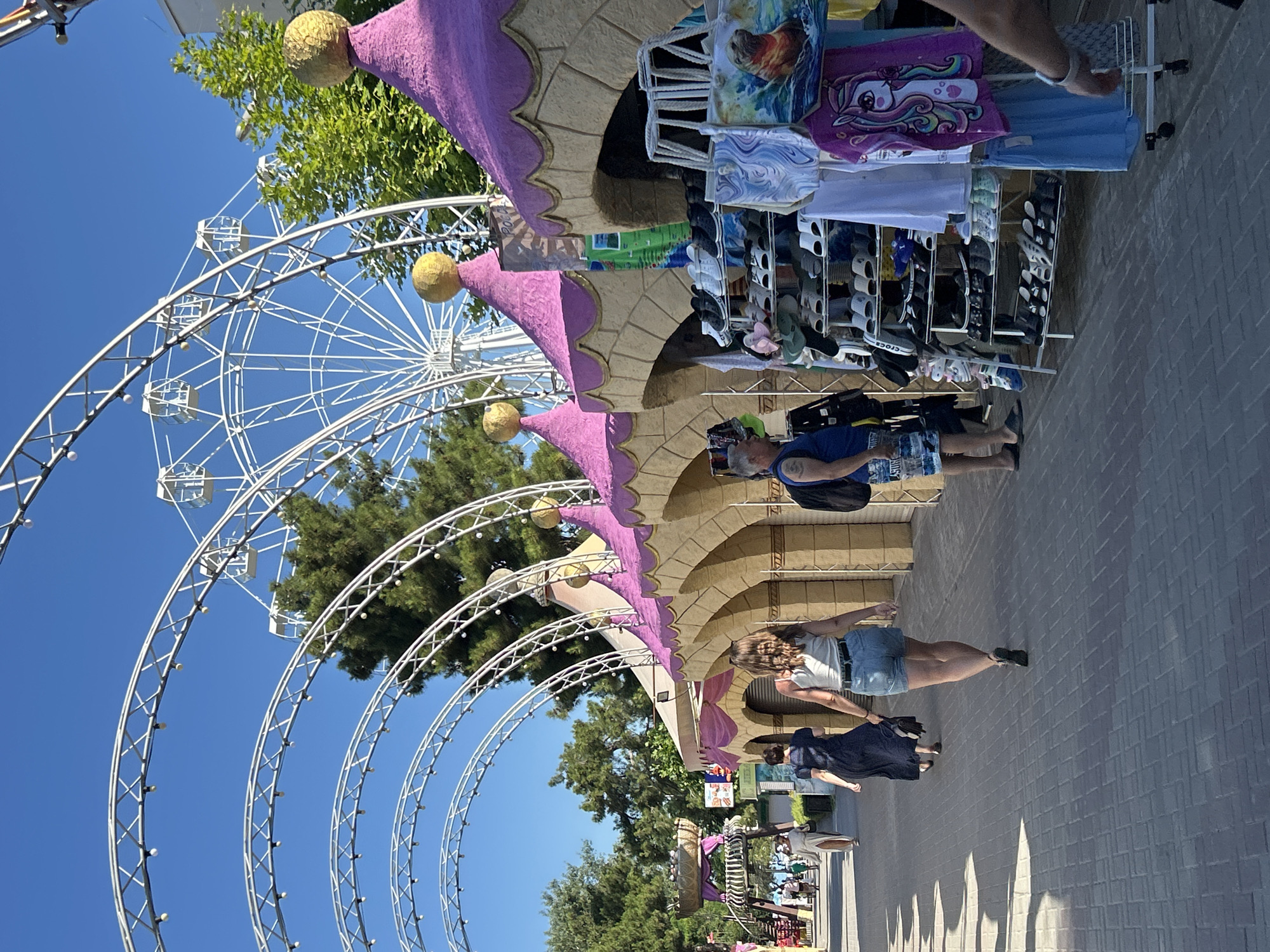 The image depicts a sunny day in Gelendzhik, Russia, with a clear blue sky overhead. In the foreground, a paved walkway is lined with arched structures and market stalls. Several people are walking along the path. To the right, a market stall displays a variety of footwear, including sandals and sneakers, as well as clothing such as t-shirts and a child's garment featuring a unicorn. Behind the stalls, carnival-like tents with purple roofs and golden spheres are visible. In the background, a large white Ferris wheel stands tall against the sky, suggesting an amusement park or entertainment area. Lush green trees frame the scene, adding to the vibrant atmosphere.