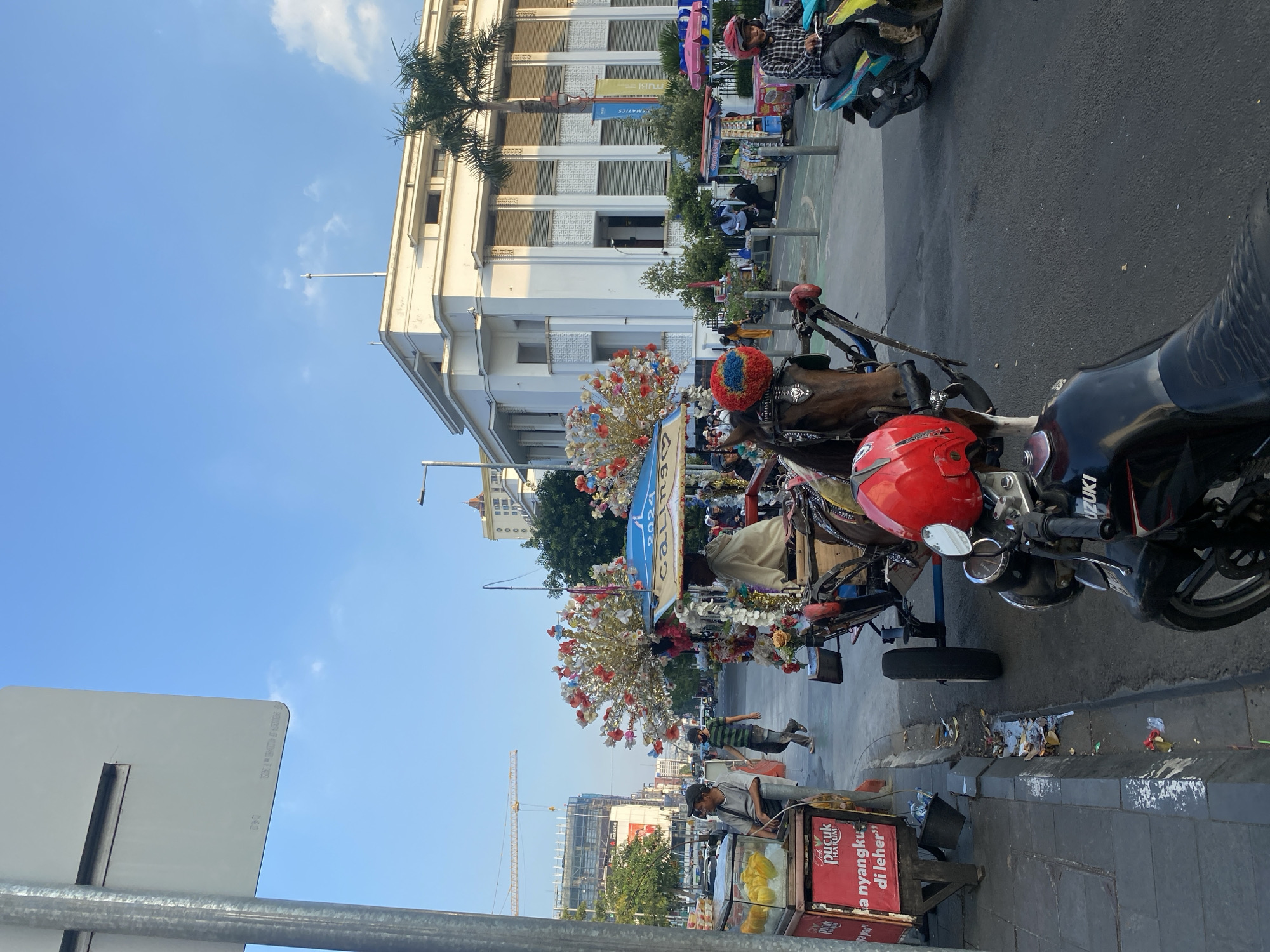 The image depicts a street scene in Kota Tua, West Jakarta, Indonesia, with a horse-drawn carriage prominently featured. The carriage, decorated with colorful flowers and artificial trees, is positioned on a cobblestone street. A brown horse is harnessed to the carriage, which has a blue canopy. A person, appearing to be the driver, is seated on the carriage.
In the foreground, a black motorcycle with a red helmet is parked. To the left, a food stall displays yellow fruits in clear containers, with red signage that reads "PUCK HARUM". A man in a gray shirt stands near the stall.
The background reveals a large, white colonial-era building with arched windows and a palm tree. Further in the distance, construction cranes and modern buildings indicate ongoing development. Several other individuals are visible walking on the sidewalk and riding motorcycles. The sky is clear blue with a few scattered clouds, and the lighting suggests it is late afternoon. The street is paved, and a curb separates the sidewalk from the road. Debris is scattered along the curb.