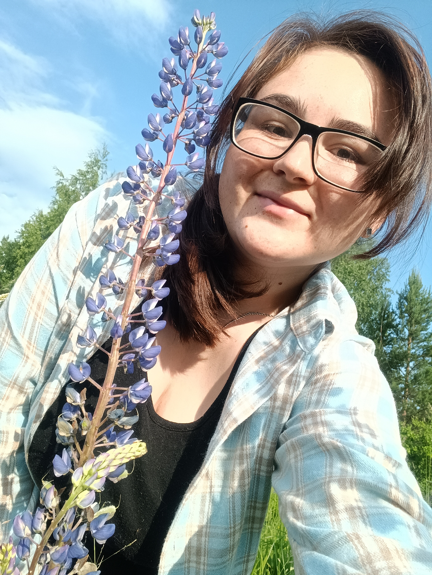 A young woman with dark hair and glasses smiles at the camera, holding a tall purple lupine flower in front of her. She is wearing a black tank top and a blue and tan plaid shirt. The background is a bright blue sky with some clouds and green trees.