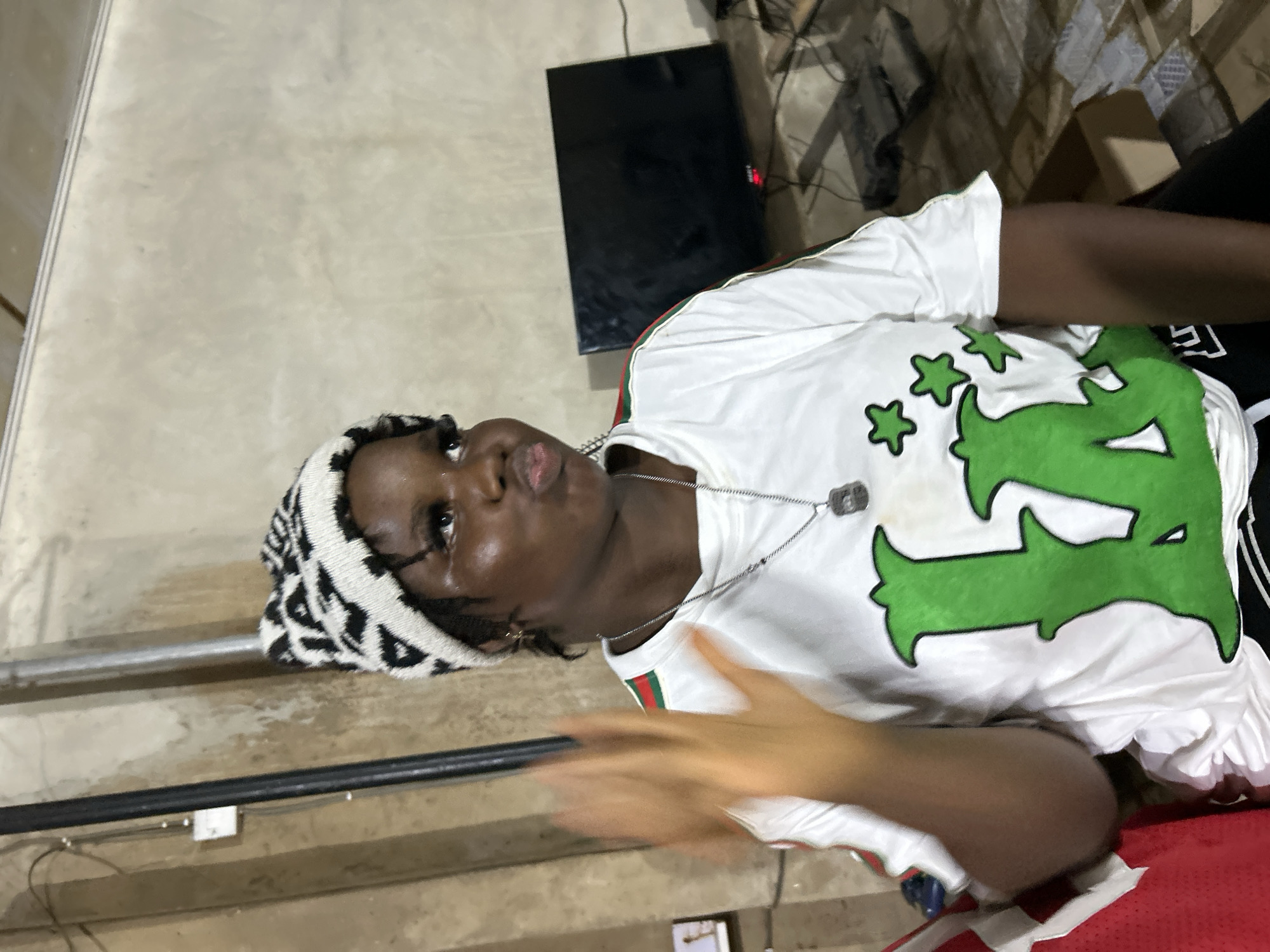 A person is looking up, wearing a black and white beanie and a white t-shirt with a large green design and a necklace. They are lying or sitting near a staircase with a black metal railing. In the background is a black flat screen TV and a cluttered area. The location is likely Ihiagwa, Nigeria. The presence of stairs and a TV suggest an indoor setting, possibly a home or apartment. A blurred hand is visible in the foreground.