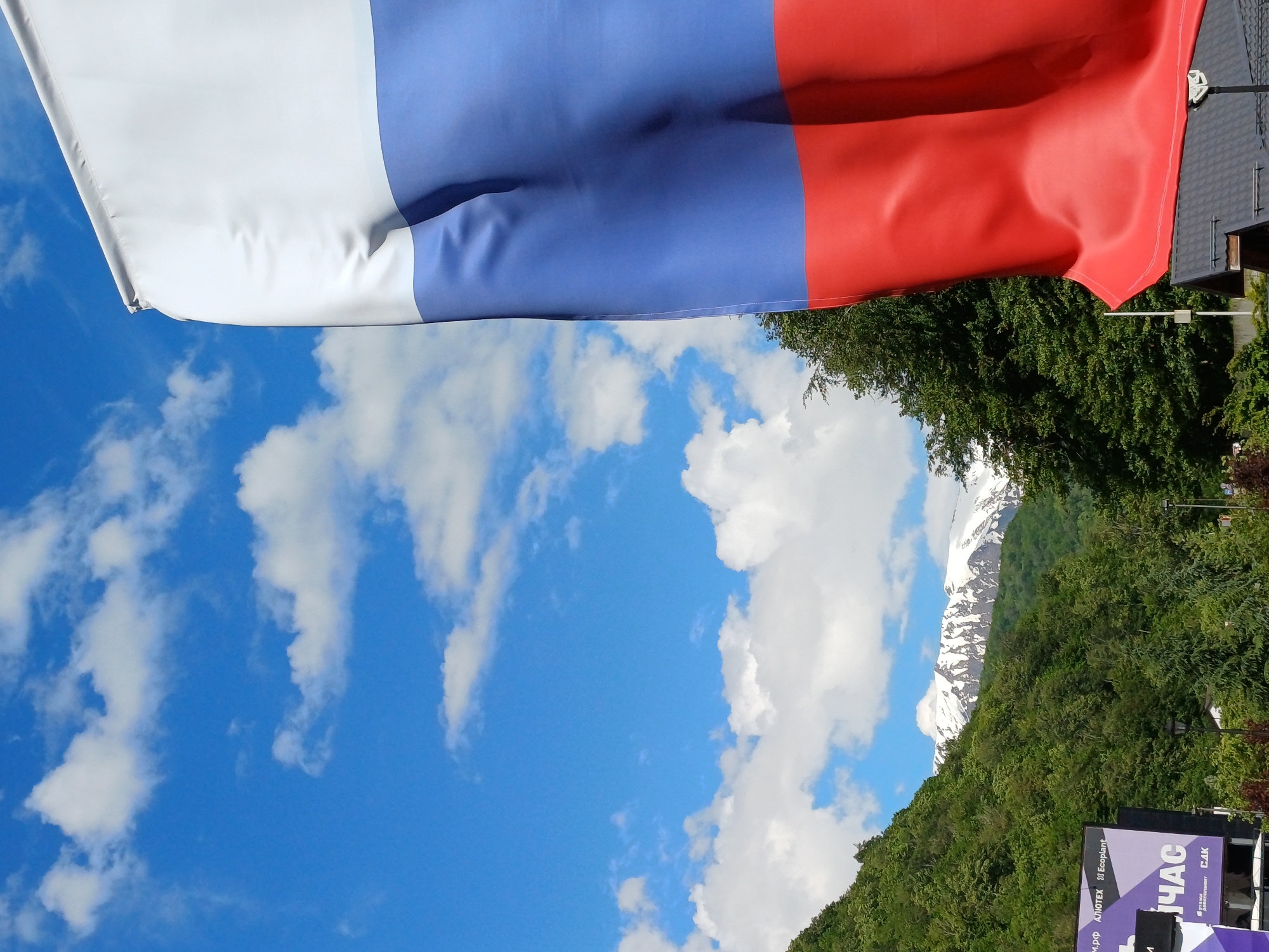 A vibrant outdoor scene in Estosadok, Russia, is depicted under bright daylight with a clear blue sky. The upper portion of the frame is dominated by a large Russian flag, unfurled horizontally. The flag displays the national colors in three horizontal stripes: white at the top, blue in the middle, and red at the bottom, showing gentle ripples from the wind.
Below the flag, a wide expanse of bright blue sky is adorned with scattered, fluffy white cumulus clouds. In the mid-ground, rolling mountain slopes are covered in dense, lush green forest. Beyond these forested hills, majestic snow-capped peaks are visible, indicating the high elevation of the mountainous terrain.
To the right side of the image, parts of a building, specifically a dark-tiled roof, and additional green foliage are partially visible. In the lower right corner, a prominent purple and black billboard displays Cyrillic text. Large white letters clearly spell "ЧАС" (CHAS), and smaller text includes "АЛЮТЕХ" (ALYUTEKH), "Экоплант" (Ekoplant), "САК" (SAK), and what appears to be ".М.РФ". The image captures a scenic, pleasant day in a mountainous resort environment.