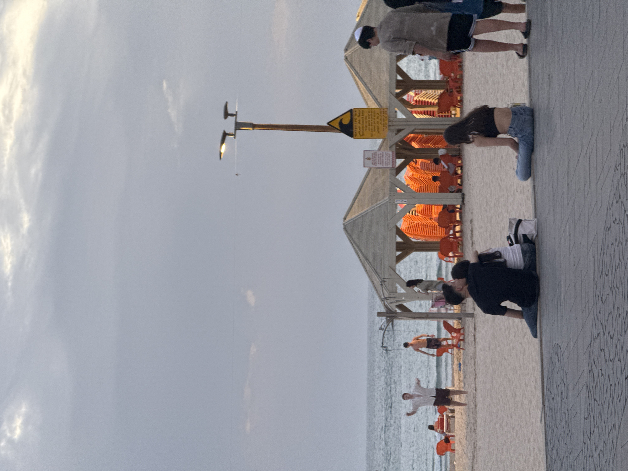 The image depicts a scene at a beach in Tel-Aviv, Israel. People are visible on the sand and the paved boardwalk area. The beach is furnished with orange beach chairs and covered structures providing shade. People are walking, standing, and seated. A lamppost stands near the boardwalk. A couple is embracing next to the path. The sky is overcast.