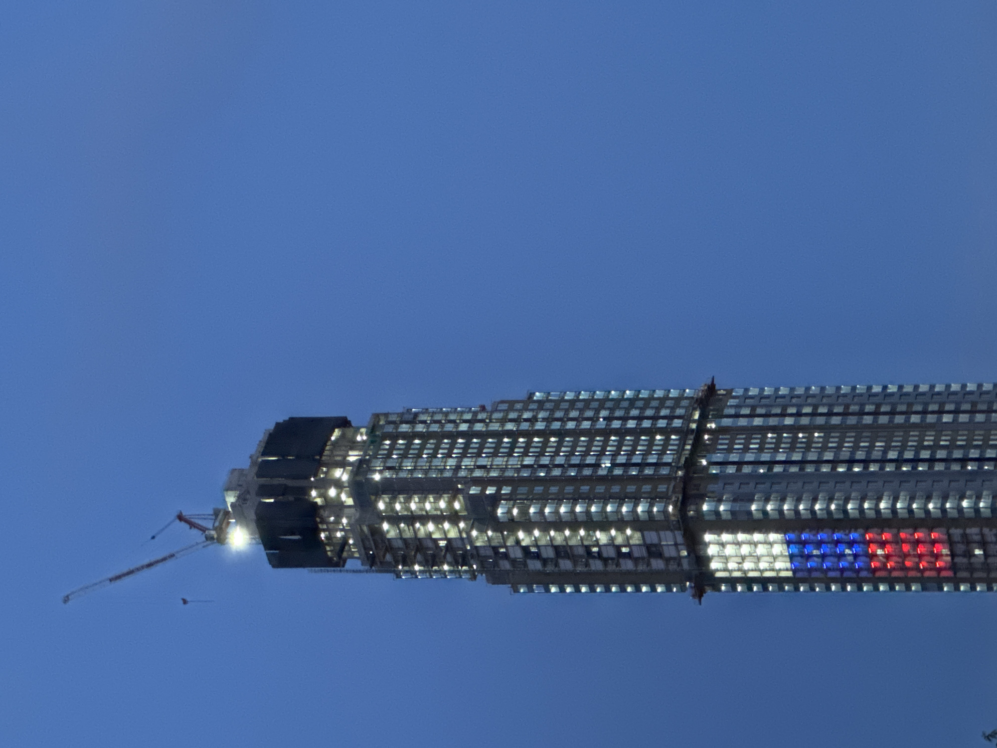 The media file presents a tall, modern skyscraper illuminated against a deep blue sky. The image is oriented with the building appearing to stretch horizontally across the frame, but upon mental rotation, it stands vertically.
The central subject is a multi-story building featuring a facade predominantly made of glass windows. Many of these windows are brightly lit from inside, creating a striking pattern of glowing rectangles across its surface. A construction crane is visible attached to the upper left side of the building, suggesting it is either still under construction or recently completed.
A particularly notable detail is a large section on the building's facade where windows are illuminated to form a distinct flag-like pattern. This pattern consists of three horizontal stripes: white at the top, blue in the middle, and red at the bottom, unequivocally resembling the national flag of Russia.
The sky is a uniform, deep periwinkle blue, indicative of either dusk or early night when artificial lights become prominent. No clouds are visible. A small, dark, elongated object, possibly a bird or a distant drone, is faintly visible in the sky near the crane.
The scene depicts a modern urban environment, specifically an architectural landmark in Moscow, Russia. The activity taking place involves the building being lit up, showcasing a national symbol. There is no visible text on the building or in the sky. The image conveys a sense of contemporary architecture and national identity.