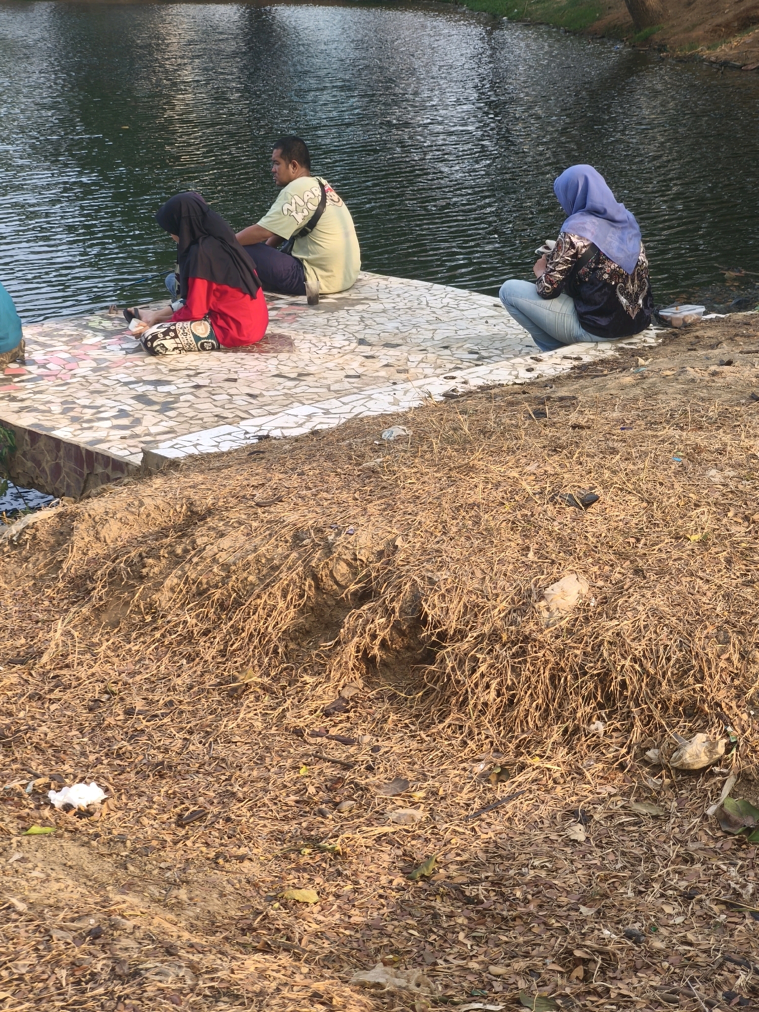 In Bekasi, Indonesia, three individuals are seated by the edge of a body of water. A man wearing a light green t-shirt and dark pants sits in the middle. To his left, a person in a red shirt and black hijab is visible. To the right, a person wearing a blue hijab, a patterned shirt, and light blue jeans is crouched down. The foreground is composed of dry, brown grass and scattered leaves, suggesting a natural or park-like setting. The water's surface is rippled, reflecting the sky. The individuals appear to be engaged in quiet activity or observation.