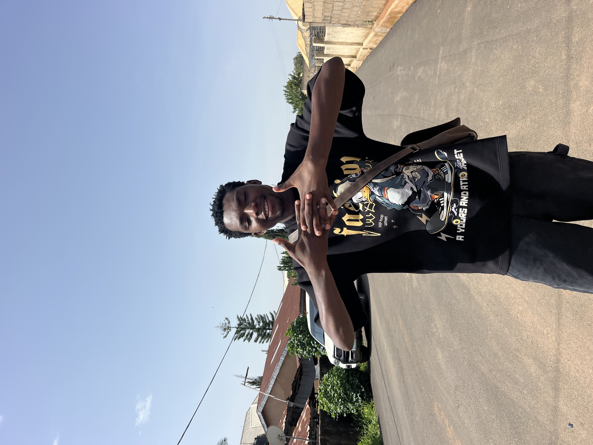 In a sunlit outdoor urban setting, a young Black male stands on an asphalt road, smiling with his eyes closed. He is wearing a black t-shirt with a graphic print of a cartoon character on a skateboard and the words "HIP-hop unset" and "A YOUNG AND ATTRACTIVE". A brown messenger bag is slung across his chest. His hands are clasped together in front of him. The road is lined with residential buildings and some vegetation. A white SUV is parked on the side of the road behind him. The sky is clear and blue.