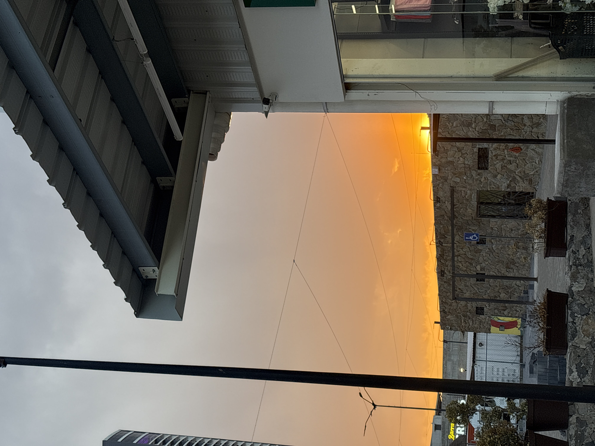The scene depicts an urban environment in San Andrés Cholula, Mexico, at sunset, characterized by a vibrant orange and yellow sky with sparse cloud cover.
In the foreground, a tall, slender black utility pole occupies the left side of the frame, extending vertically. To its immediate left, a portion of a modern high-rise building with a curved, multi-windowed facade is visible. Below the utility pole, several dark brown planters containing dry or sparse vegetation rest upon a low stone wall.
To the right, the corner of a commercial building features a light-colored facade with large glass display windows. A green section is visible above the window, and a white security camera is mounted on the upper corner, facing outwards. An overhead awning with a corrugated metal roof is supported by exposed steel beams, including a prominent beige I-beam. Through the glass, items resembling clothing (pink/red fabric) and white floral arrangements are discernible.
The midground reveals a paved area leading to a low structure clad in rough-hewn stone, featuring dark metal uprights and a visible doorway or entrance. A blue international accessibility symbol (wheelchair icon) is affixed to this stone structure. Behind it, a chain-link fence, a white shipping container, and a wall bearing an abstract mural in yellow, red, and blue are visible. Another streetlight pole with multiple lamp fixtures is positioned further back on the left, silhouetted against the sky. Overhead wires traverse the expanse of the sky.
In the background on the left, a building with partial yellow neon signage spelling "Barra RRES" is visible. No individuals are present, and no actions or interactions are depicted within the scene.