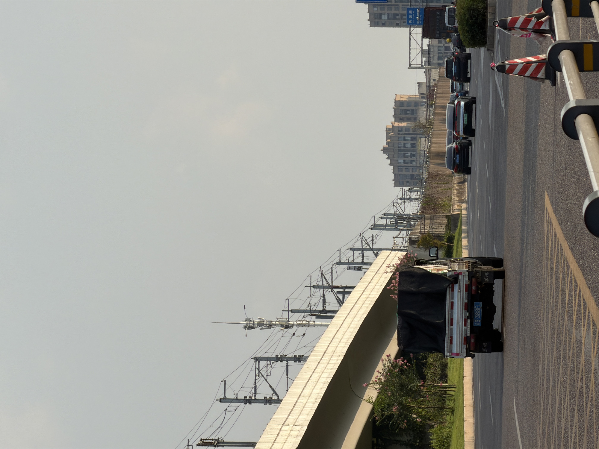 In Jiangjin, China, a flatbed truck with a black tarp covering its cargo drives away from the viewer on a multi-lane road. The truck's license plate reads "B-388EL". Behind it, several cars, including a black sedan and a silver sedan, are also traveling on the road. In the background, tall buildings and an elevated railway structure dominate the urban landscape. The sky is clear and pale. The road is marked with yellow and white lines. Traffic cones and guardrails are visible along the side of the road.