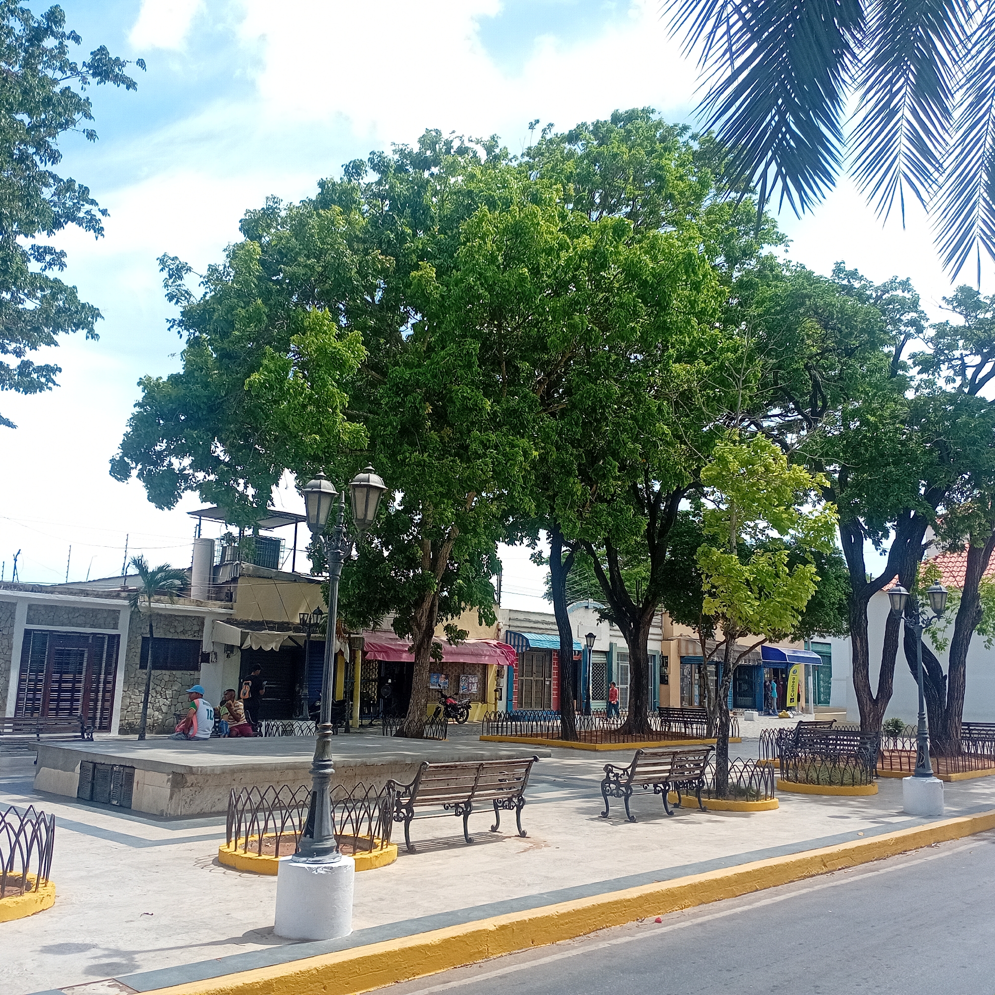 The image shows a plaza in an urban setting, likely a town square. The plaza features concrete paving with a yellow curb bordering a road. Several trees provide shade, with benches placed beneath them and around the edges of raised planter areas. Vintage-style lampposts are positioned around the plaza.
In the background, buildings line the perimeter of the square, including shops with awnings. Two people are seated on a raised platform in the plaza. The sky is blue with scattered clouds. The overall environment appears to be a public space for relaxation and commerce.