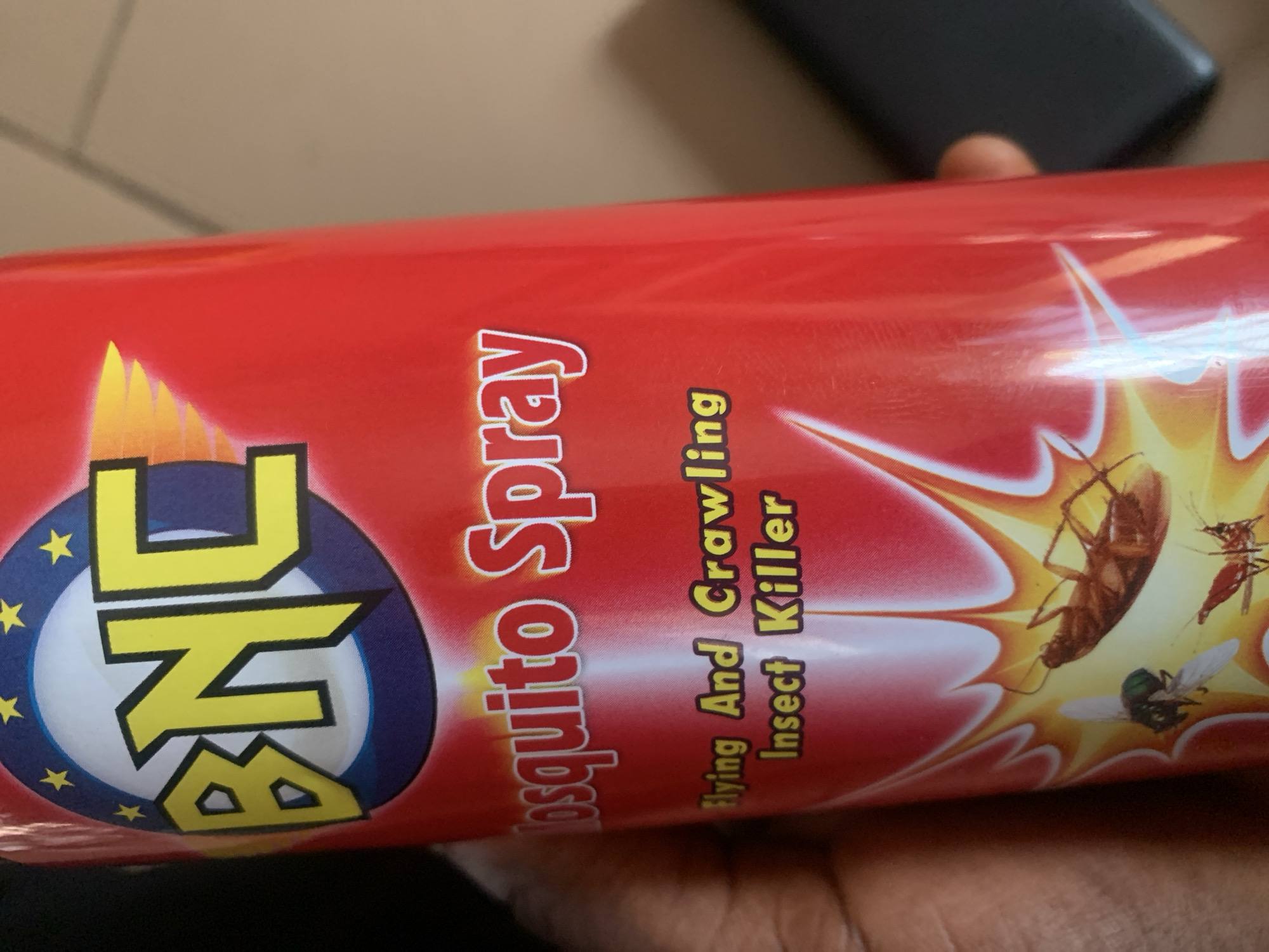 A can of BNC Mosquito Spray is held by a person's hand. The can is primarily red with yellow lettering that reads "BNC" in a stylized font at the top, surrounded by a blue circle and yellow stars. Below this, "Mosquito Spray" is written in white lettering with red outlines. Further down, "Flying And Crawling Insect Killer" is printed in yellow text. The lower portion of the can features graphics of insects, including a cockroach, a fly, and what appears to be a mosquito or similar biting insect, all depicted within a yellow burst design. The background is blurred, but suggests an indoor environment with a tiled surface and a dark object, possibly a phone, to the right. The image is taken in Jauro Yinu, Nigeria, as indicated by the context provided.