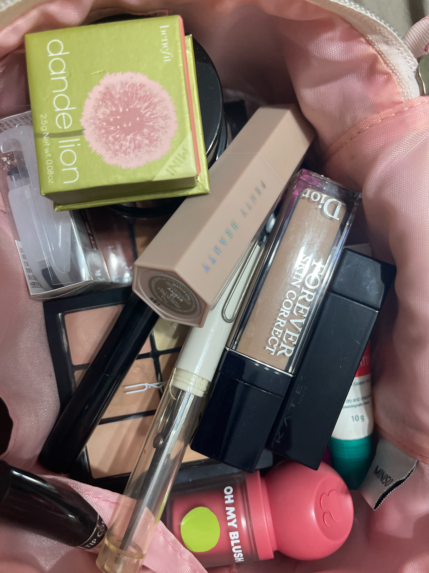 This image shows a collection of makeup items inside a pink makeup bag. The makeup items include a Benefit "Dandelion" blush, a Fenty Beauty foundation stick, a Dior "Forever Skin Correct" concealer, a black eyeliner pencil, and a pink cream blush. A small tube labeled "MINISO" is also visible. The lighting is dim, suggesting an indoor setting. The image does not contain any information about the city of Ipoh, Malaysia, nor does it depict any people or actions.