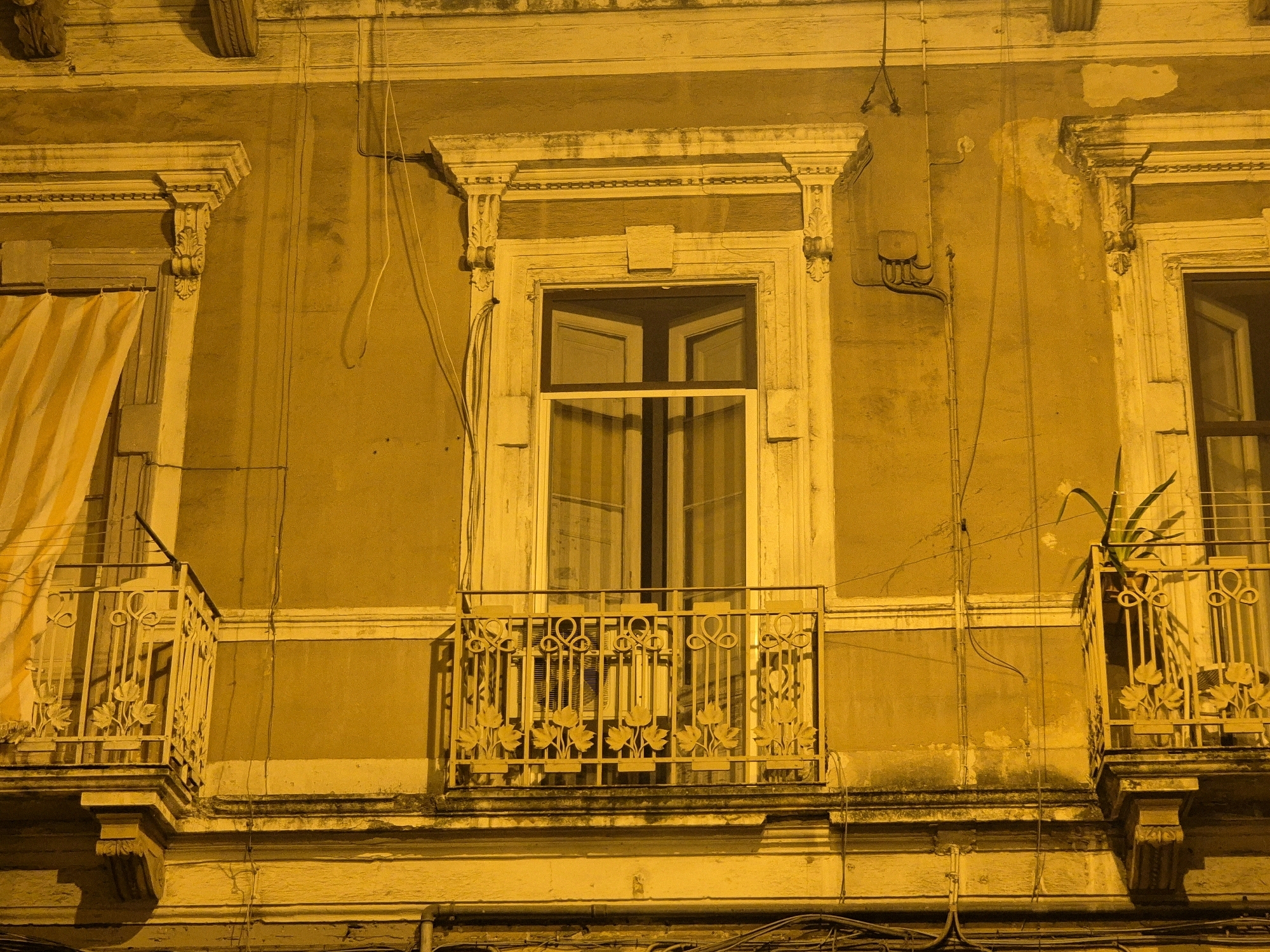 The image depicts a building facade in Taranto, Italy, at night, illuminated with a yellowish-orange light, likely from street lamps or artificial interior lighting. The building features classic architectural elements, including ornate window frames with decorative moldings and corbels supporting balconies.
The facade itself appears to be plaster, showing signs of age and some discoloration or peeling in certain areas. There are multiple windows visible; the central window is large and appears to be a double-hung or casement window with visible internal blinds or curtains showing vertical stripes. To the left of the central window, a portion of a striped curtain or fabric is visible hanging outside the frame, suggesting a window opening behind it, possibly covered.
Two balconies with decorative wrought-iron railings are present. The railings have an elaborate design featuring scrollwork and stylized leaf motifs. The balcony on the left is partially obscured by the striped fabric. On the right balcony, a potted plant with green foliage is visible, with a long stem reaching upwards.
Electrical conduits and wiring are visible running vertically and horizontally across the facade, attached to the wall in several places. A small electrical box with several conduits entering it is also present on the wall to the left of the central window.
The lower portion of the image shows the underside of the balconies and the building's base, with more electrical wiring and what appears to be a rougher texture of the building material. The overall impression is of an older building in an urban environment, possibly a residential or commercial structure, with visible signs of utility infrastructure. No people are present in the scene.