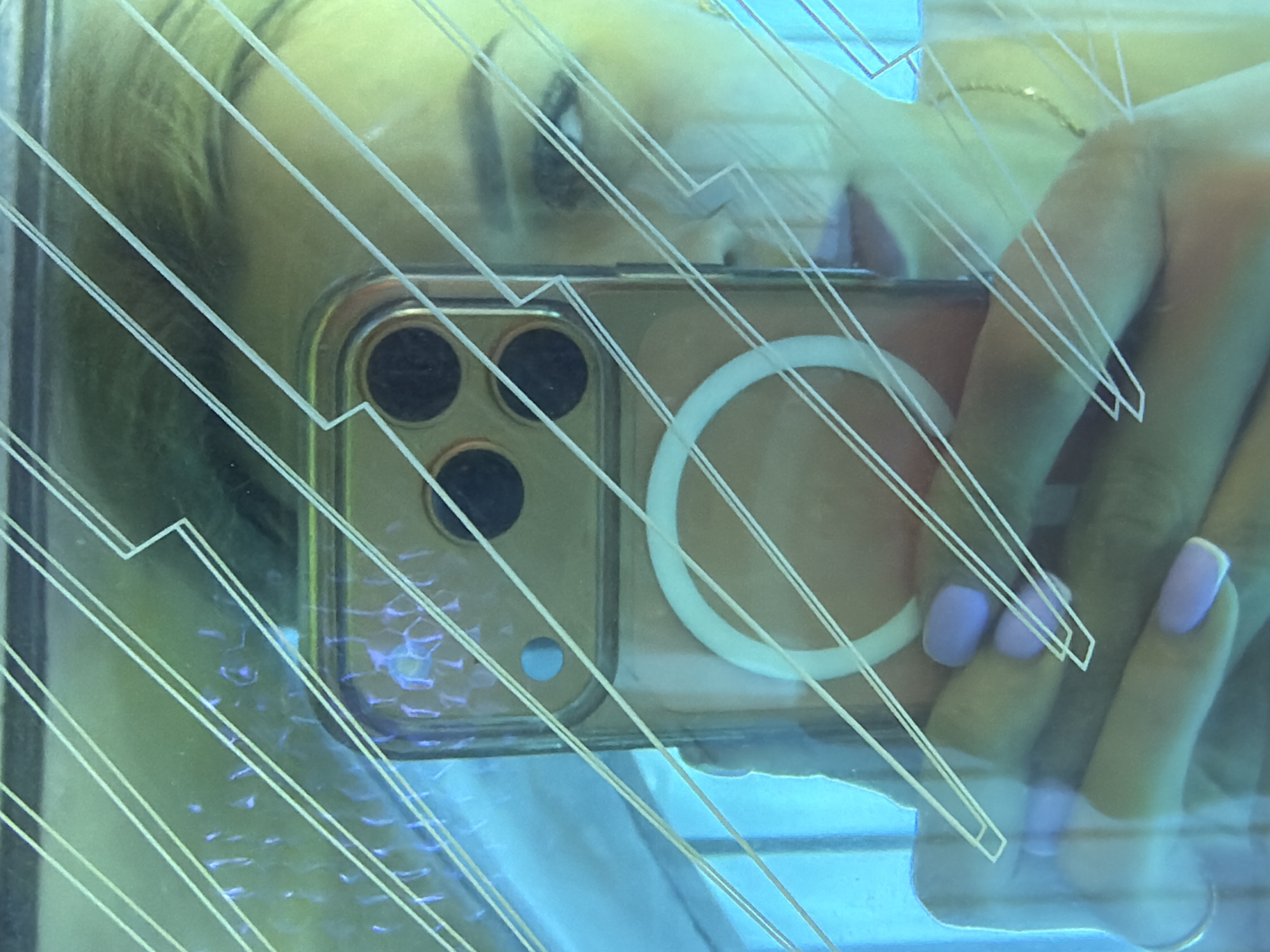 A selfie is taken in what appears to be a tanning bed. The subject, a woman, is holding a phone with a rose gold case and a white ring attached to it. The phone's camera is visible, as are three circular lenses. The woman's face is reflected in the phone screen, and she is wearing light purple nail polish. The tanning bed has a reflective surface with a pattern of parallel lines. The lighting is predominantly blue and green. There is no discernible location in the city of Kyiv, Ukraine.