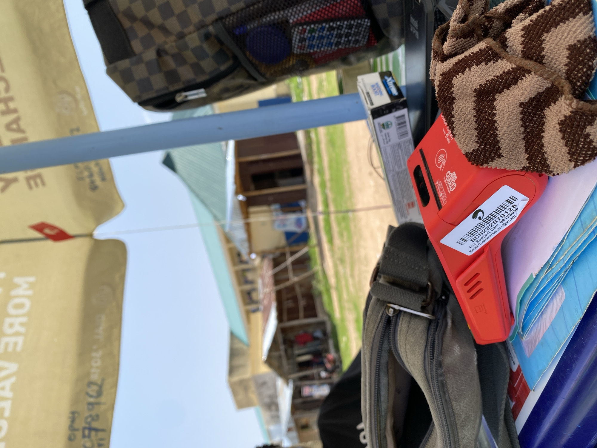 This is a close-up, eye-level, outdoor shot in Jalingo, Nigeria, featuring a red point-of-sale device with a label that reads "SC0222070128 For Support Call: 09..." and an email address. Beside the device is a striped brown and tan cloth and a green canvas bag with its zipper partially open. In the background, there is a checkered brown and black backpack and the blurred outlines of buildings and trees under a blue sky. The image appears to have been taken during daylight.