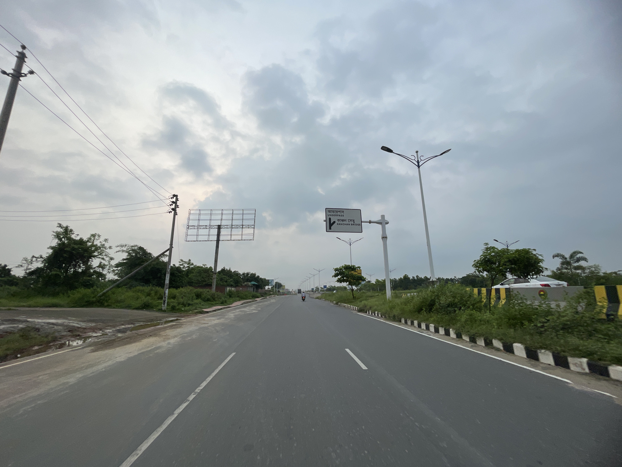 The image depicts a road in Purbachal, Bangladesh under a cloudy sky. The road is asphalt, marked with white lane lines and flanked by black and white curbs. On the left side of the road, there is a puddle of water near overgrown vegetation and utility poles. A billboard frame stands to the left, and a road sign indicating "Underpass" and "Kanchan Bridge" is visible in the middle of the road. Streetlights line the road on both sides. In the distance, a single motorcyclist is visible. The environment is characterized by green vegetation and an overcast sky.