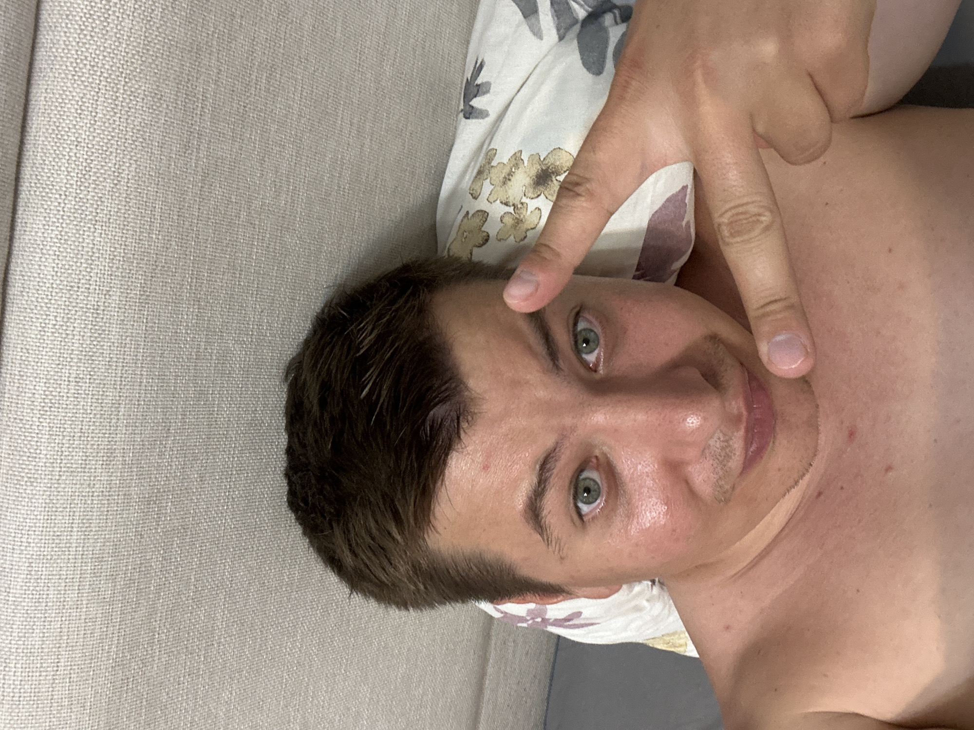 A close-up, overhead selfie shows a shirtless person lying on a bed, with their head resting on a pillow that has a floral pattern. The person has short, dark brown hair and light green eyes. Their face is illuminated, with a slight sheen of sweat on their forehead and cheeks. They are looking directly at the camera and have their right hand raised, with two fingers extended in a peace sign. The background consists of a textured, light-colored surface, likely the headboard of a bed. Based on the visual cues and common imagery associated with Budva, Montenegro, this scene likely depicts a hotel room or a private residence in the city, possibly taken during a warm period.