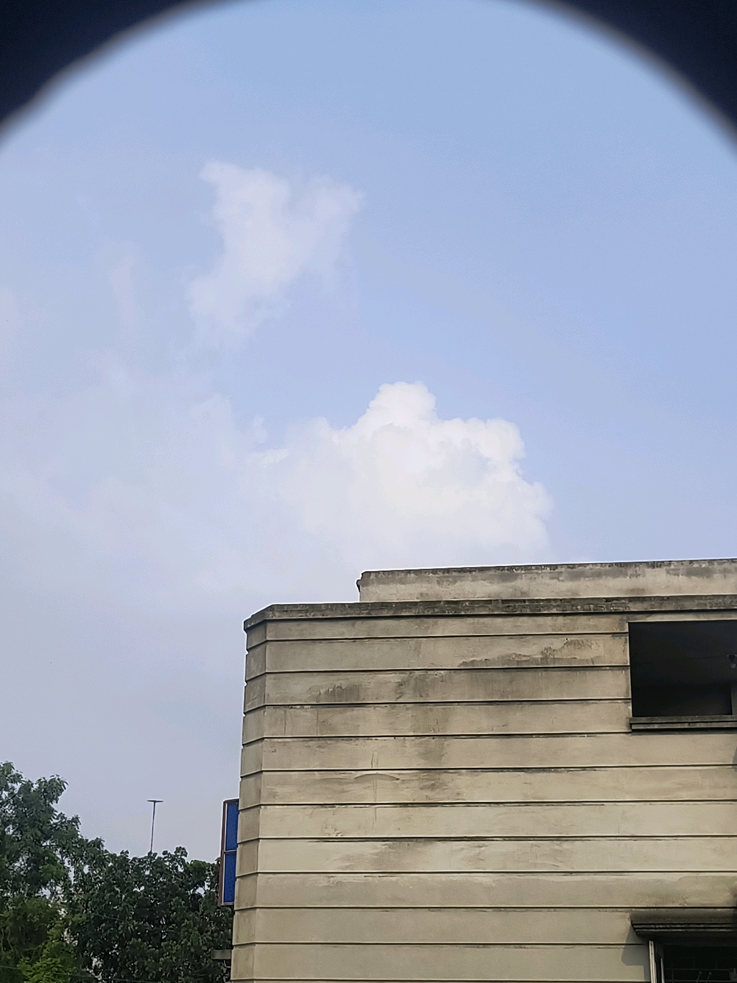 This image, taken in Beldanga, India, captures a portion of a building with horizontal concrete siding against a light blue sky with scattered white clouds. The building's facade is weathered, showing signs of age and staining. To the left of the building, the upper branches of lush green trees are visible. A small, dark opening, possibly a window or vent, is present on the upper right side of the building. The top edge of the image is obscured by a dark, curved border, suggesting it might have been taken through a circular lens or aperture. The overall scene conveys an urban environment with hints of natural elements.