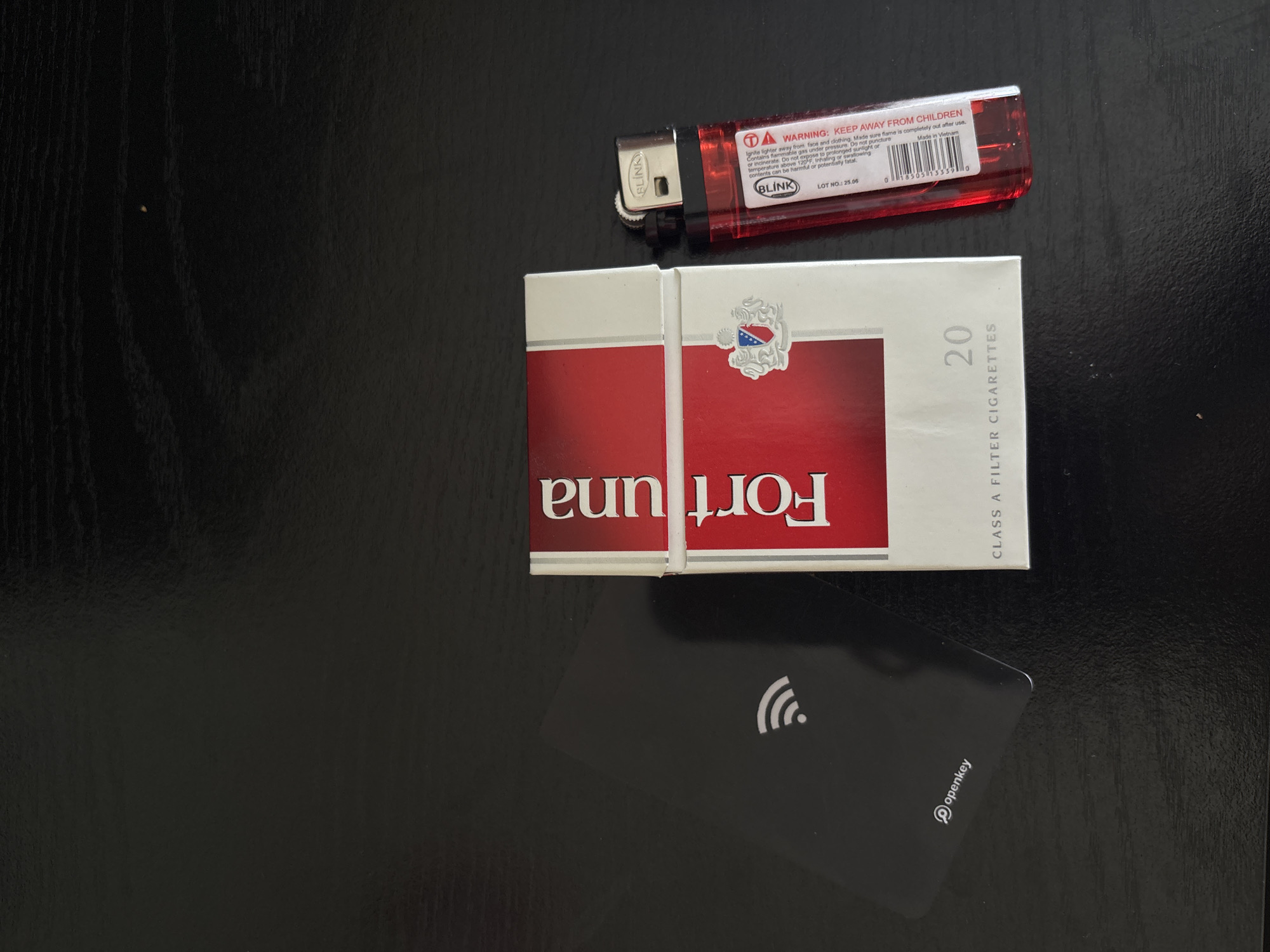An overhead, close-up shot shows a black table with a pack of Fortuna cigarettes and a red Blink lighter. The pack of Fortuna cigarettes is vertical and to the left of the lighter. The pack is mostly white with a red stripe that has "Fortuna" written in white lettering. To the left of the cigarettes, there is a black access card with a wifi symbol on it, and the word "openkey" in white lettering near the bottom. The red lighter is to the right of the cigarettes, and is lying on its side. The lighter has "Blink" written on it, and a warning label that says "WARNING: KEEP AWAY FROM CHILDREN." The image appears to be taken indoors. The location in San Francisco, United States of America is not visible in the image.