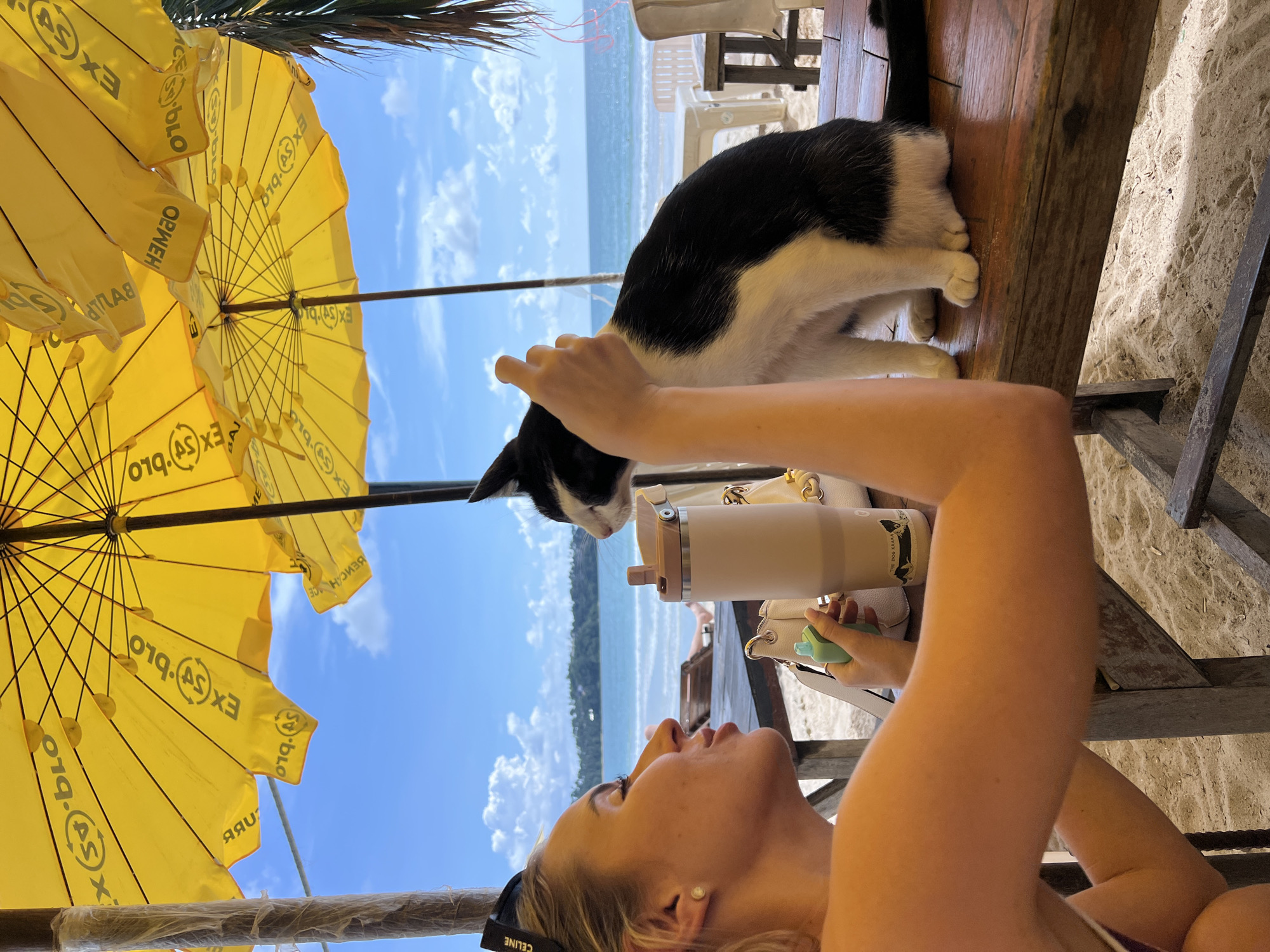 A black and white cat sits on a wooden table on a sandy beach. A young woman is to the left of the cat, leaning in to pet it. She is wearing a black headband with "CELINE" written on it. She is holding a green vape in her right hand. A beige insulated cup and a white purse are on the table. In the background, there is a blue ocean, white clouds, and several yellow umbrellas providing shade. The location is a beach, likely a resort or cafe given the table and chairs.
