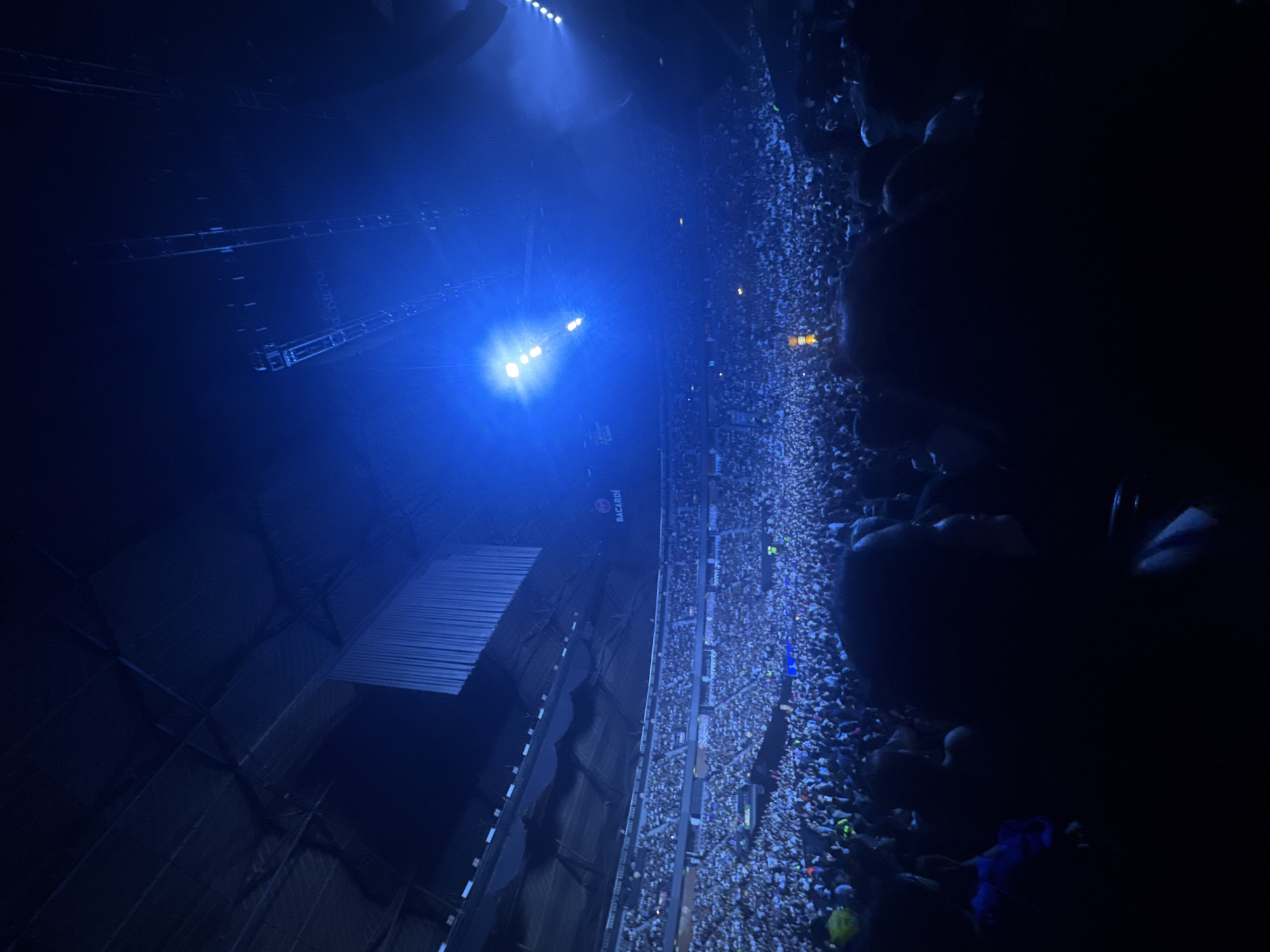 The image shows a large crowd of people gathered at a concert venue in Mexico City, Mexico. The venue is illuminated with blue lighting. There are stage lights and scaffolding visible above the crowd. A Bacardi sign is visible on a wall. The crowd appears dense, with many individuals visible in the frame. Some people are looking towards the stage, while others are looking at their phones. The photo seems to have been taken from an elevated position, possibly a balcony or upper level within the venue.