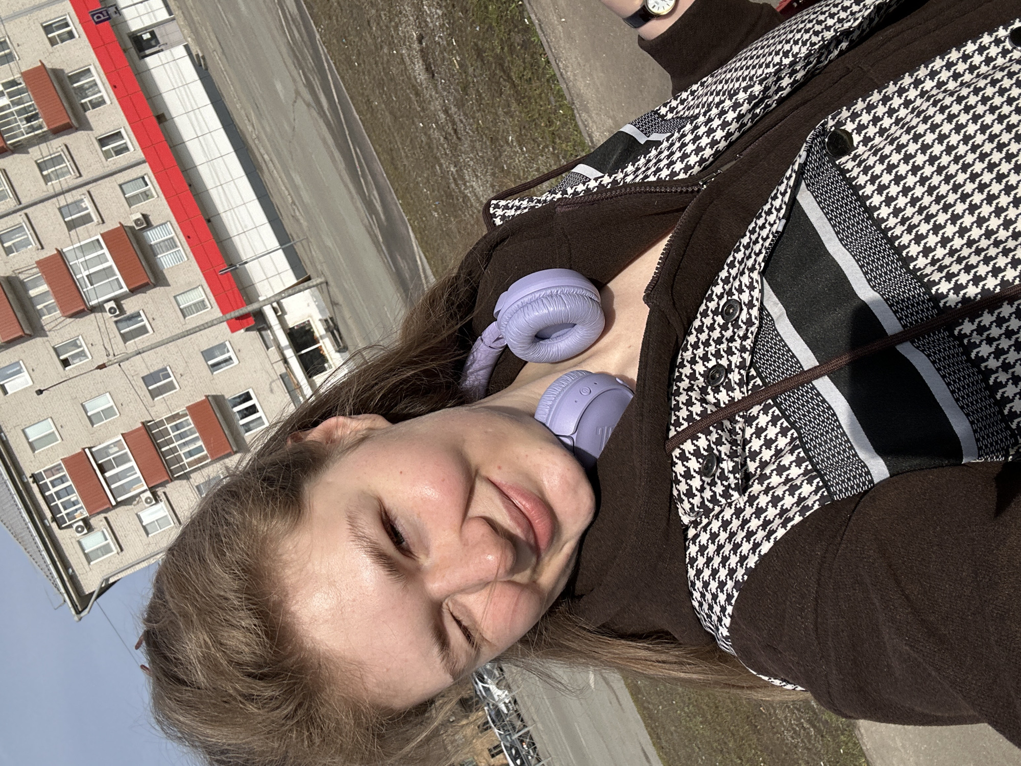 A woman is seen in a selfie. She is wearing a brown jacket with a black and white houndstooth pattern. Lavender colored headphones rest around her neck. A building is in the background, with white and gray walls and red roofs. The scene is in the city of Kazan, Russia.