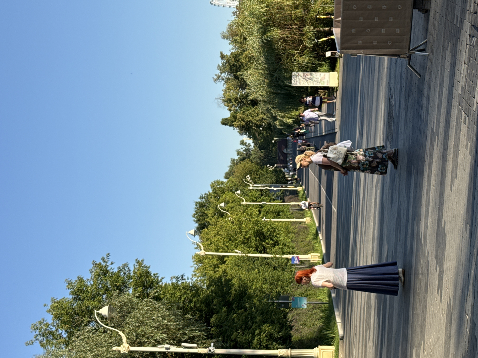 The image depicts a bright, sunny day on a wide pedestrian promenade, likely in Moscow, Russia, given the characteristic lampposts and architectural style. The promenade is bordered by lush green trees on the left and a street with light traffic on the right. Several people are visible, strolling and cycling.
In the foreground, two women stand facing away from the camera. The woman on the left has bright red hair and wears a white, sheer top with a long, flowing dark blue skirt. The woman on the right, wearing a straw hat, is dressed in a floral print maxi dress and carries a handbag. They appear to be standing still, perhaps conversing or observing the surroundings.
Further down the promenade, more people are walking, and a cyclist is seen on the street. Streetlights, typical of urban environments, line the promenade. The ground is paved with cobblestones or patterned concrete. In the background, a Ferris wheel can be seen on the right, indicating a recreational area. On the far right, a temporary barrier or signage structure is present. The overall scene conveys a peaceful, active urban atmosphere.