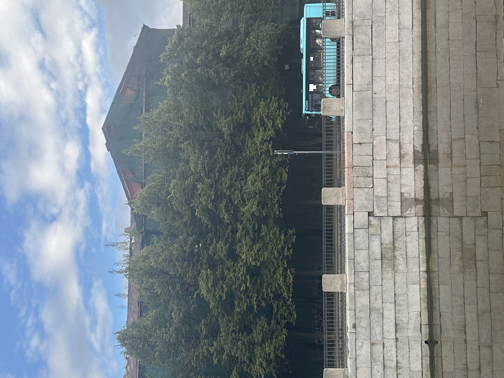 The image depicts a turquoise bus traveling along a street bordered by a stone embankment and a canal in Saint Petersburg, Russia. The bus, with "VOLGABUS" visible on its side, is seen from its left flank, revealing several passengers inside. Behind the street and trees, a large building under renovation is partially visible, covered in green netting. The sky above is partly cloudy. The canal water reflects the stone embankment.