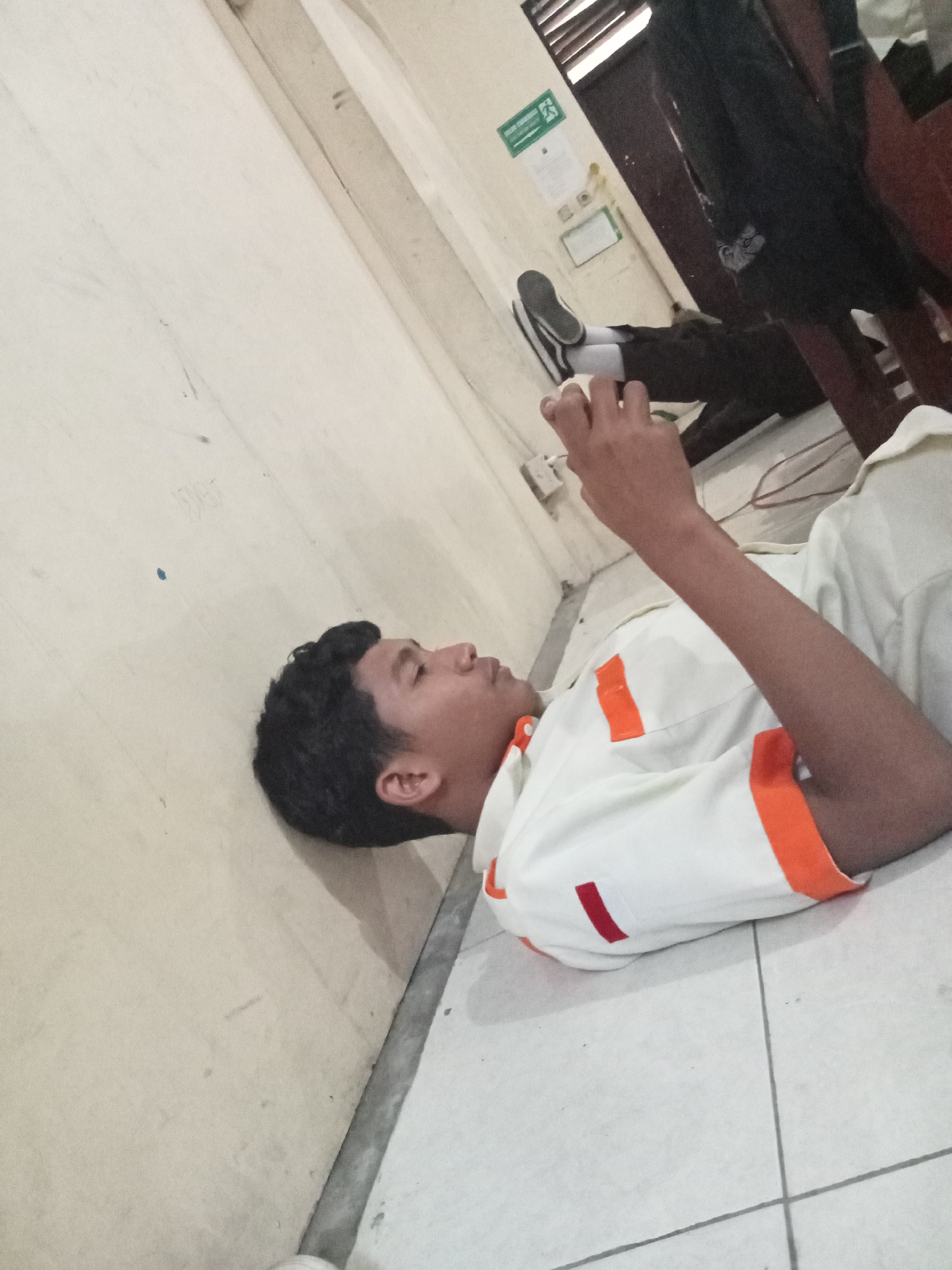 The image depicts a young male lying on his back on a tiled floor, leaning against a light-colored wall. He is wearing a white shirt with orange accents on the collar, sleeves, and chest pocket, and dark pants. His right arm is extended, and he appears to be holding a mobile phone. His head is turned to the left, and his eyes are looking downwards. In the background, a dark figure is partially visible, seated on a wooden chair. A small green safety sign is mounted on the wall. The lighting suggests an indoor setting, possibly a classroom or common area. The overall scene appears to be within a building in Secang, Indonesia, given the context of the provided image location.