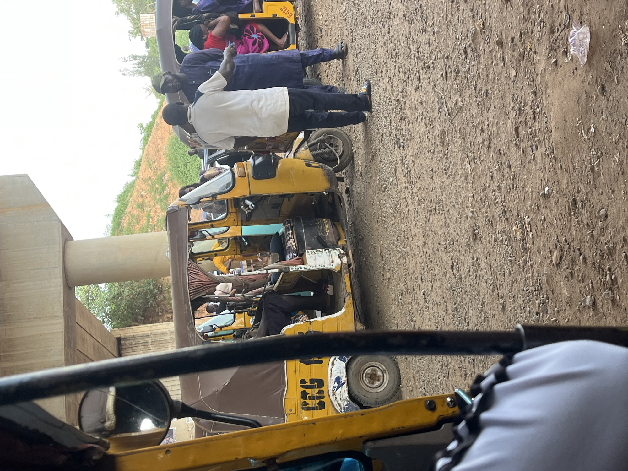 The image shows a scene in Galadimawa Village, Nigeria, likely at a transport hub or roadside. In the foreground, the front of a yellow auto-rickshaw is visible, with a black bar obscuring part of the view. To the right of this auto-rickshaw, another similar yellow auto-rickshaw is parked, with its side door open. Inside this second auto-rickshaw, several passengers are seated, including a person in a pink and blue patterned skirt and a red top. Two men are standing near the open door of this auto-rickshaw, one wearing a white t-shirt and dark trousers, and the other in a dark blue outfit and a green cap. They appear to be interacting, possibly discussing transport. Behind these auto-rickshaws, a road or pathway is visible, with a concrete overpass structure overhead, supported by large pillars. The ground is covered with gravel and dirt. In the background, there is a hillside with sparse vegetation. A second auto-rickshaw, also yellow, is partially visible further to the right, with passengers inside. The overall impression is of a busy, informal transportation area.