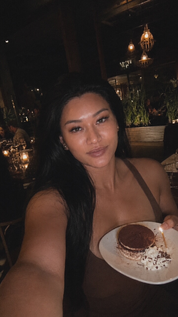A medium shot selfie captures a woman with long dark hair and light skin, wearing a brown top, as she holds a plate with a small dessert topped with chocolate shavings and whipped cream, and a lit candle. The dessert is a round, layered pastry, possibly a tiramisu. The woman is looking towards the camera with a neutral expression. She is in an indoor dimly lit environment that appears to be a restaurant, with other patrons and decorative lighting visible in the background. The image is taken at night.