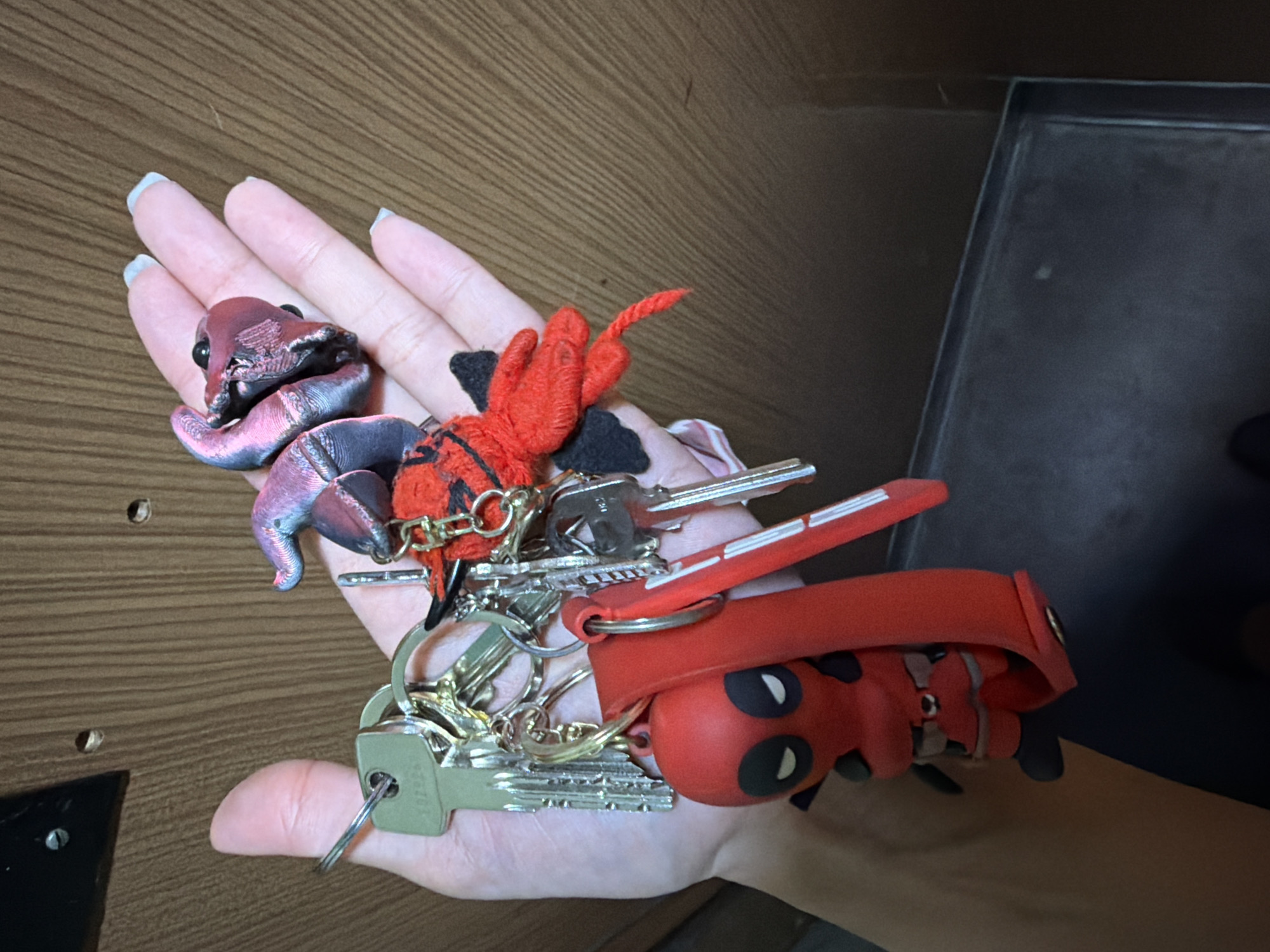 A hand with light skin and light blue painted fingernails is holding a set of keys with several keychains attached. One keychain is a red figure of Deadpool. Another is a red devil plush toy. A third keychain is a shiny, iridescent fabric resembling a sea creature with dark eyes. The background appears to be a wooden surface, possibly a door or wall, with vertical grain patterns. The lighting suggests an indoor environment. The image does not provide any information about a location in Sofia, Bulgaria.