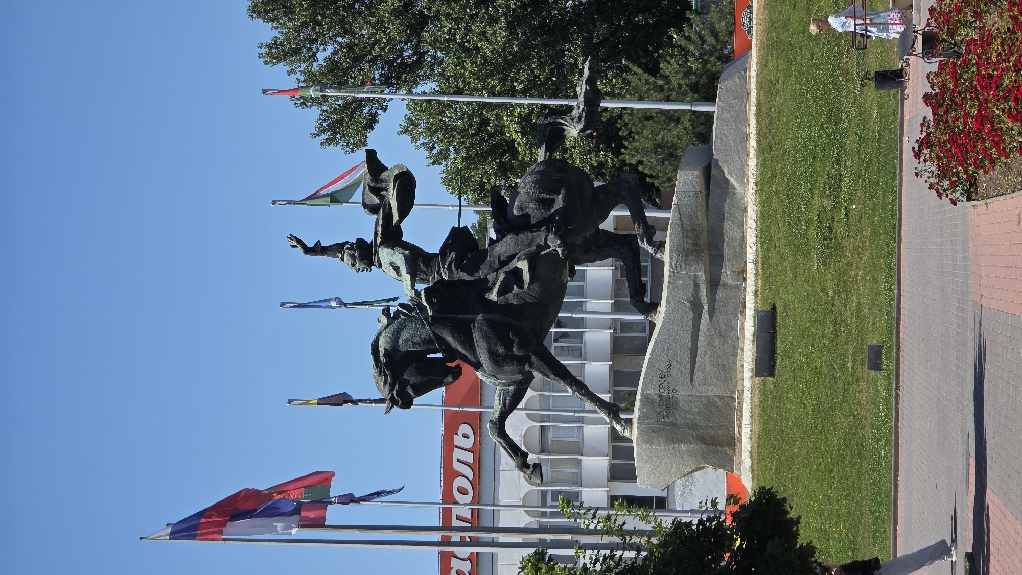 The image depicts the Alexander Suvorov monument in Tiraspol, Moldova, set against a clear blue sky. The monument features a bronze equestrian statue of a man in military attire, with his arm raised. The statue stands on a large stone pedestal inscribed with Cyrillic text, likely indicating its dedication and the year of its erection.
In the background, a white building with red signage is visible, suggesting a commercial or public establishment. Several flags are displayed on poles to the left and right of the monument; one prominently features the colors of the Russian flag. Lush green grass covers the ground around the monument, and trees provide a backdrop.
In the foreground, a paved walkway with red and grey brickwork is visible. A woman in a light-colored dress is walking on the right side of the walkway, carrying a bag. On the left side, a person wearing light grey pants and a dark top is walking away from the camera. Red flowers are planted in a bed along the edge of the walkway on the right. The overall scene is a sunny day in a public urban space.