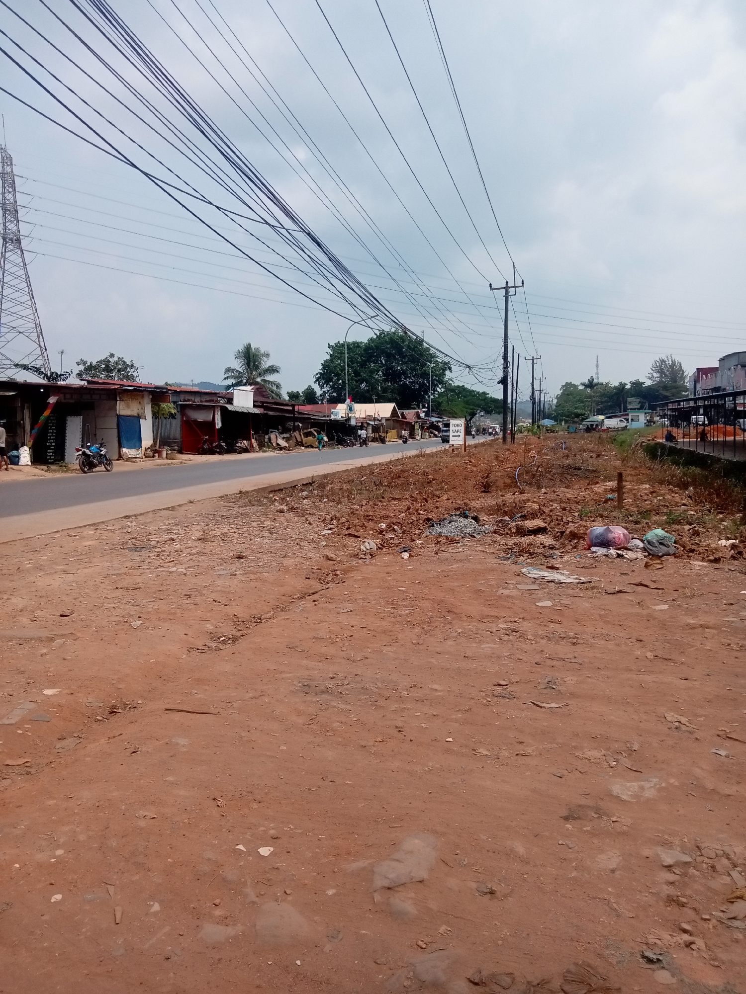 The image depicts a roadside scene in Batam, Indonesia, characterized by a dusty, unpaved foreground and a paved road lined with simple buildings and overhead power lines. The foreground is a reddish-brown dirt surface, unevenly distributed with small rocks, debris, and scattered refuse, including plastic bags and what appears to be paper or fabric.
Adjacent to the road, on the left side of the frame, are a series of small, makeshift structures, likely shops or residences, with corrugated metal roofs. A blue and white sign with the text "TOKO VAPE" and an arrow pointing to the left is visible. Several motorcycles are parked along the road in front of these buildings. A few people are seen walking or standing near the shops.
On the right side of the road, the environment is a mix of overgrown vegetation and more debris, including piled-up garbage bags. A few trees and palm trees are scattered throughout the scene, and in the distance, more buildings and vegetation can be observed. Overhead, a complex network of power lines crisscrosses the sky, supported by utility poles and a tall transmission tower on the far left. The sky is overcast with clouds. The overall impression is of a developing or somewhat neglected urban or peri-urban area.