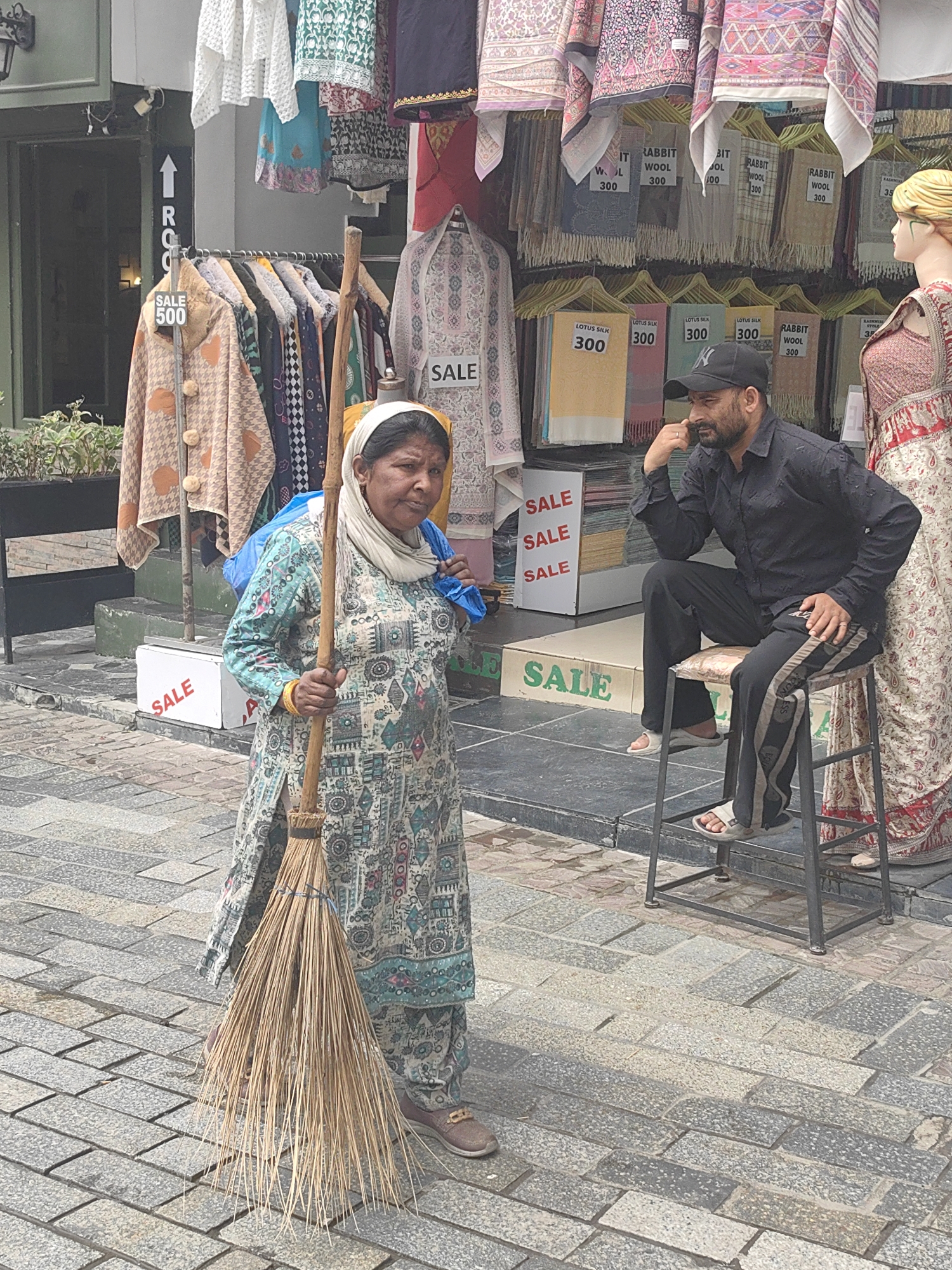 A woman stands on a cobblestone street holding a broom. She is wearing a light-colored headscarf and a long, patterned dress. Behind her, a store displays clothing and textiles, with several "SALE" signs visible. To her right, a man in a black shirt and pants sits on a stool, looking to his left. A mannequin in a patterned dress stands to the far right. The scene appears to be a marketplace or shopping area in Manali, India.