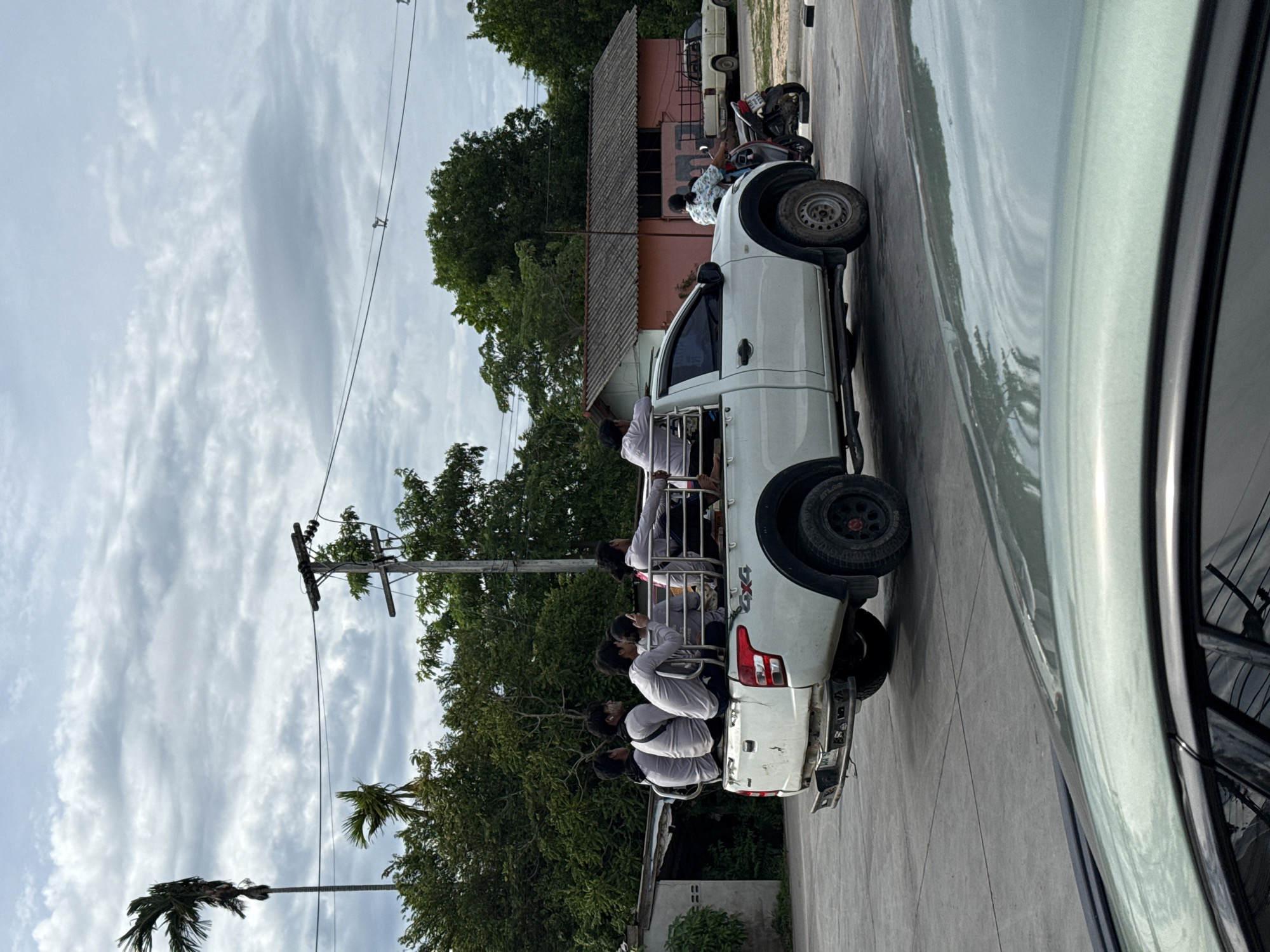 A white pickup truck with a modified bed, containing six young males wearing light grey shirts, is parked on a paved road in the city of Ko Samui District, Thailand. The individuals are seated on a bench and appear to be engaged in conversation. The truck is positioned on the right side of the frame. In the background, a single-story pink building with a dark grey roof is visible, along with lush green trees and a cloudy sky. To the right of the truck, a motorcycle is parked. Another white pickup truck is parked further in the background. The scene suggests a casual outdoor setting with a focus on transportation and local life.