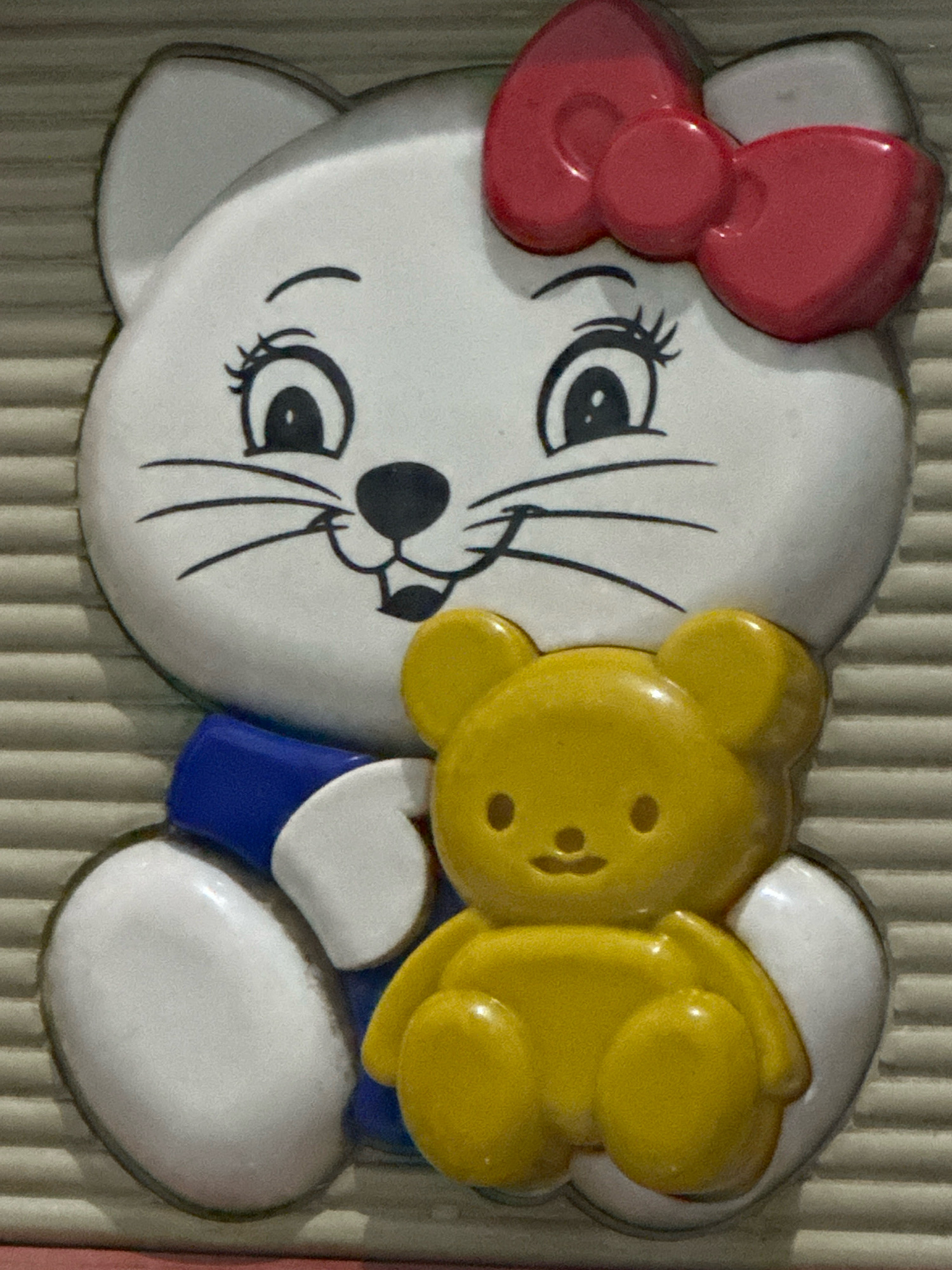 The image shows a close-up, eye-level shot of a wall-mounted decoration featuring a white cat character with a red bow on its head, and a yellow teddy bear character being held by the cat. The cat has large black eyes with eyelashes, a black nose, and thin black whiskers. It is wearing a blue collar or shirt. The teddy bear is small and yellow, with two black dots for eyes and a small black mouth. Both characters are embossed or raised from the surface of the decoration, which appears to be made of plastic or a similar material. The background is a ribbed, light-colored surface, possibly a wall or a textured panel. The lighting is somewhat diffused, creating soft shadows that highlight the three-dimensional nature of the decoration. There are no people present in the image. The object is a decorative piece, likely for a child's room or a themed area, and its exact location within the city of Binjai, Indonesia, cannot be determined from the visual information provided.