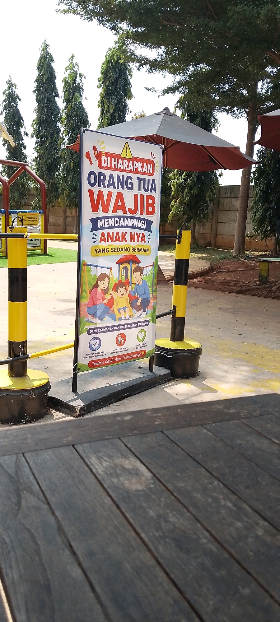 The image depicts an outdoor scene, likely at a children's play area, with a prominent sign in Indonesian. The sign, a vertical banner with colorful illustrations, is the central subject. It features a family with two parents and a child playing, alongside text that translates to "Parents are expected to accompany their children while playing." The sign also includes icons and text emphasizing safety and the importance of supervision.
The sign is propped up by a dark base and flanked by two bollards, one on each side, painted with alternating yellow and black stripes. These bollards appear to be part of a barrier or entryway. In the background, there are tall, slender trees with dense foliage, and a concrete wall. Behind the sign and bollards, a children's playground structure with slides and climbing frames is visible. There is also an open umbrella-like structure, suggesting an area for shade.
The ground surface is a combination of light-colored concrete and a grassy area. The foreground is dominated by a wooden surface, possibly a deck or table, with visible planks and a weathered appearance. The lighting suggests it is daytime, with bright sunlight casting shadows.
The context of "Sriamur, Indonesia" suggests this scene is located in a public outdoor space within that area, likely a park, playground, or recreational facility designed for families and children. The overall impression is a well-maintained area focused on child safety and family enjoyment.