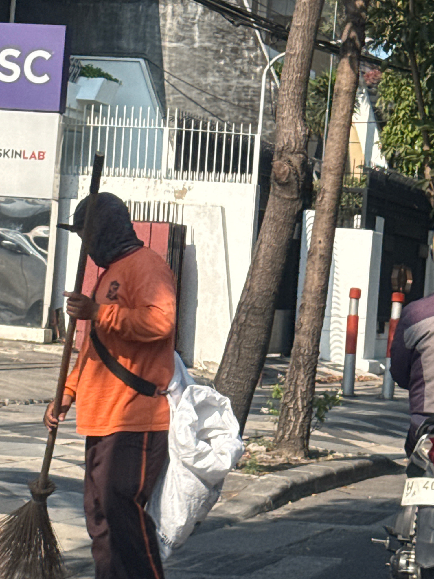 A person wearing an orange long-sleeved shirt and dark pants, with their head covered by a dark cloth, is standing on a sidewalk holding a broom. A large white bag is slung over their shoulder, and a black strap is visible across their chest. Behind the person, a white fence with vertical bars separates the sidewalk from a building. To the left, a purple sign with white letters that read "SC" is visible. Further to the left, a sign with red letters that read "SKINLAB" is also present. Two trees with rough bark stand in the foreground, with their trunks obscuring parts of the background. In the background, a white building with a concrete facade and a dark roof can be seen. To the right of the frame, the rear of a motorcycle with a license plate displaying "W 40" is visible. The scene appears to be an outdoor urban environment with paved roads and sidewalks.