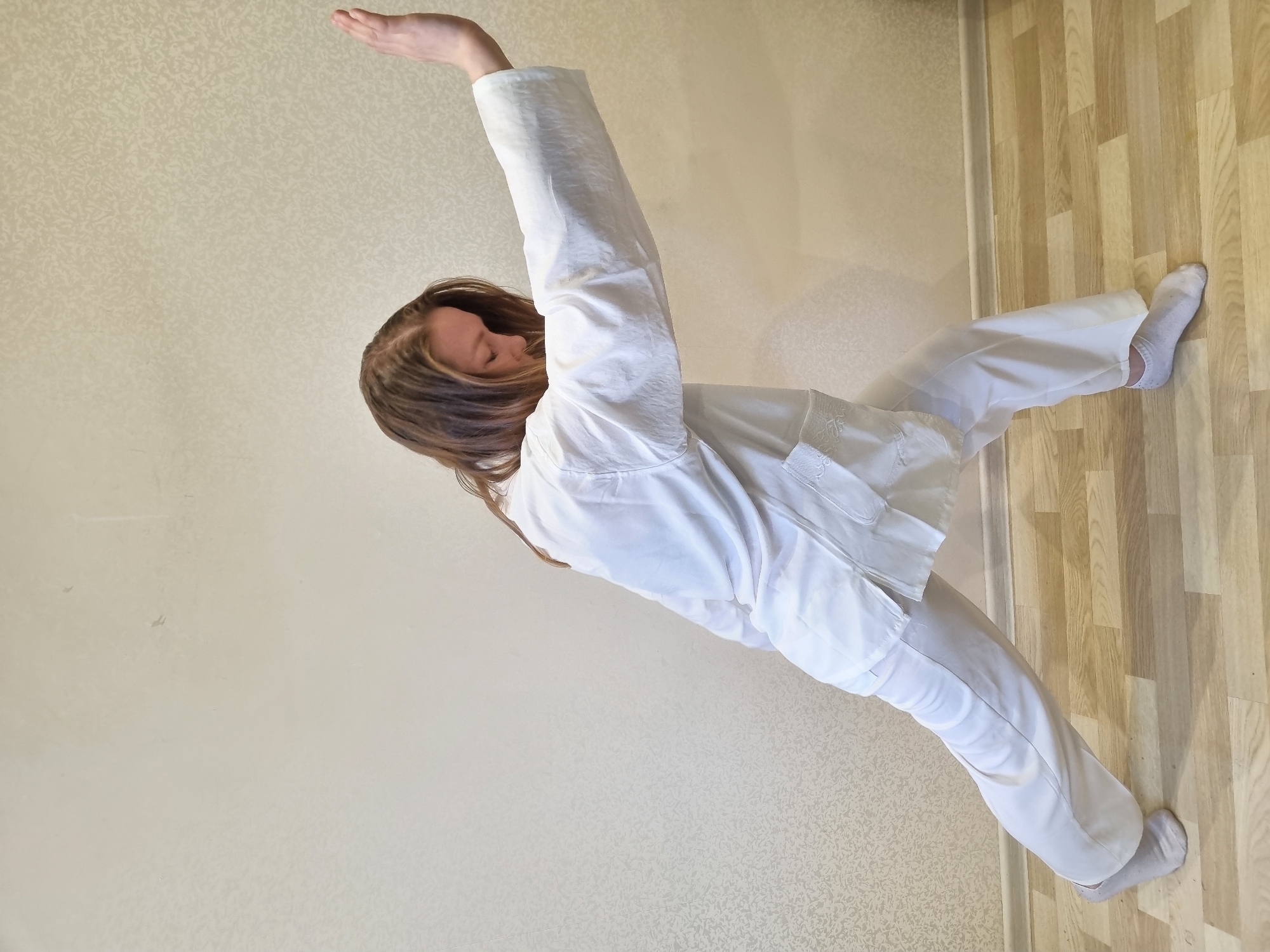 A woman is indoors in a room in Chaikovsky, Russia. She is wearing white clothing and socks and is extending one arm above her head while standing on one leg. The flooring is light-colored wood laminate, and the wall is off-white with a subtle pattern.