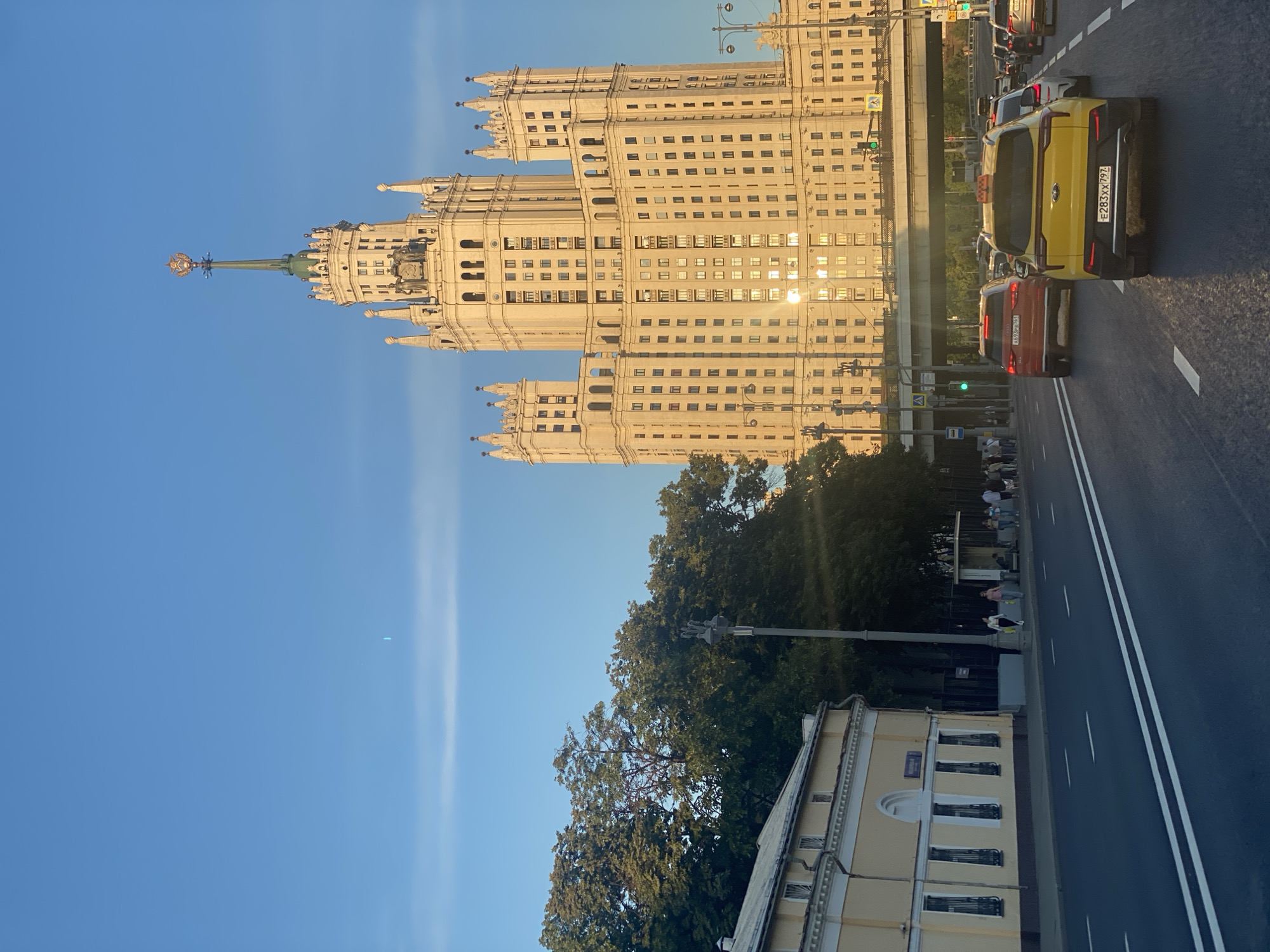 The image captures a view of the Kotelnicheskaya Embankment Building, a prominent Stalinist skyscraper located in Moscow, Russia. The building, bathed in the golden light of what appears to be late afternoon or early evening, dominates the skyline with its imposing stature and ornate architecture, including spires and a prominent star emblem at its apex. In the foreground, a multi-lane road is visible with several cars, including a distinctive yellow taxi, moving in traffic. The road is lined with white markings. To the left of the skyscraper, a lower, pale yellow building with barred windows and an arched entrance is visible, with a section of green trees behind it. People are seen walking on the sidewalk near the yellow building and a bus stop. The sky is a clear blue with a thin, wispy cloud formation near the horizon and a faint streak of light, possibly a contrail, higher up. The scene is characteristic of an urban setting in Moscow, with the skyscraper serving as a recognizable landmark.