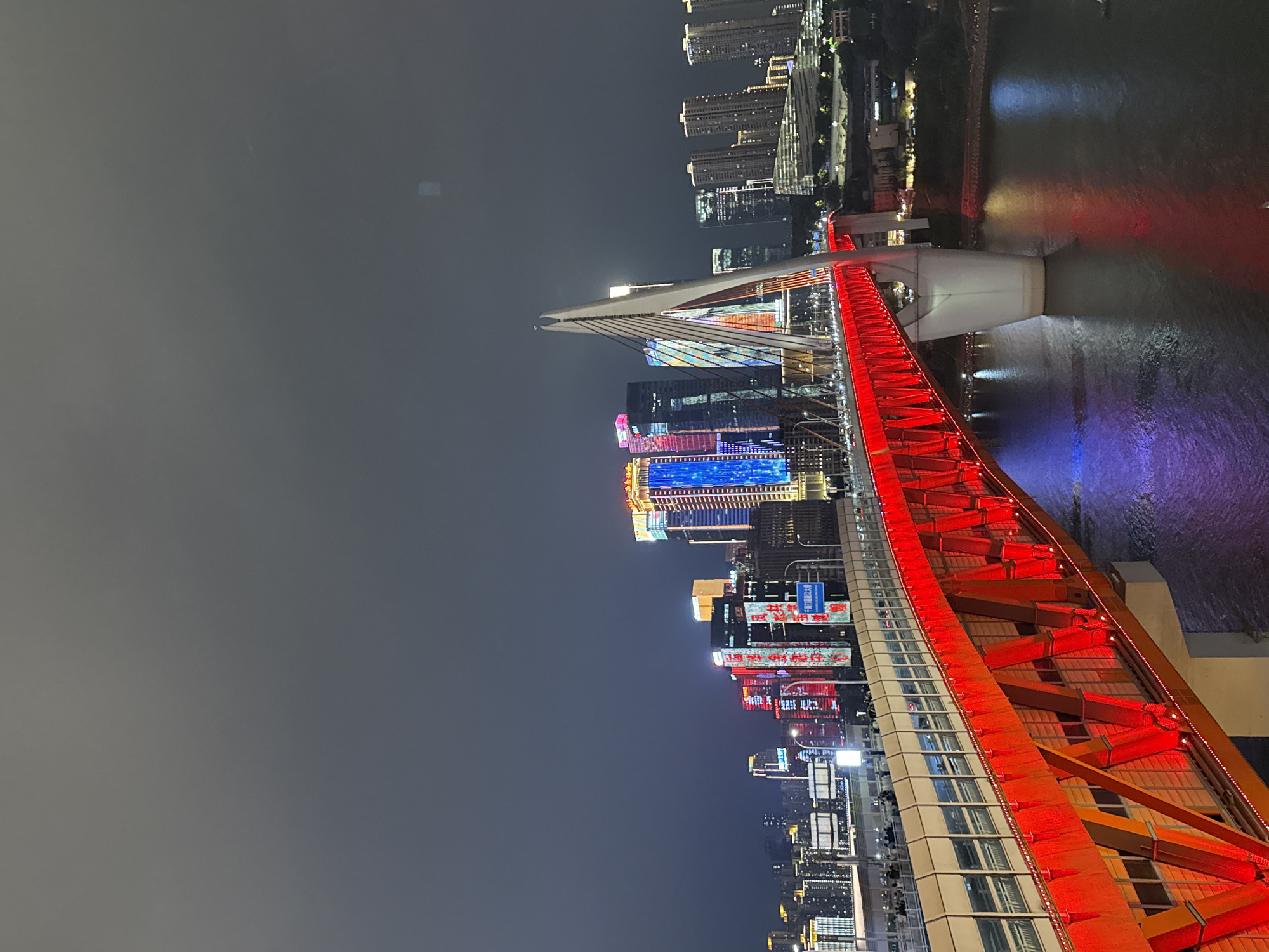 The image is a night shot of the red-painted steel structure of the Qiansimen Bridge in Chongqing, China, situated within the Yuzhong District. The bridge is illuminated, with the red light reflecting in the river below. In the background, a cityscape is visible with brightly lit skyscrapers, their reflections also visible in the water. To the left is a dark sky.