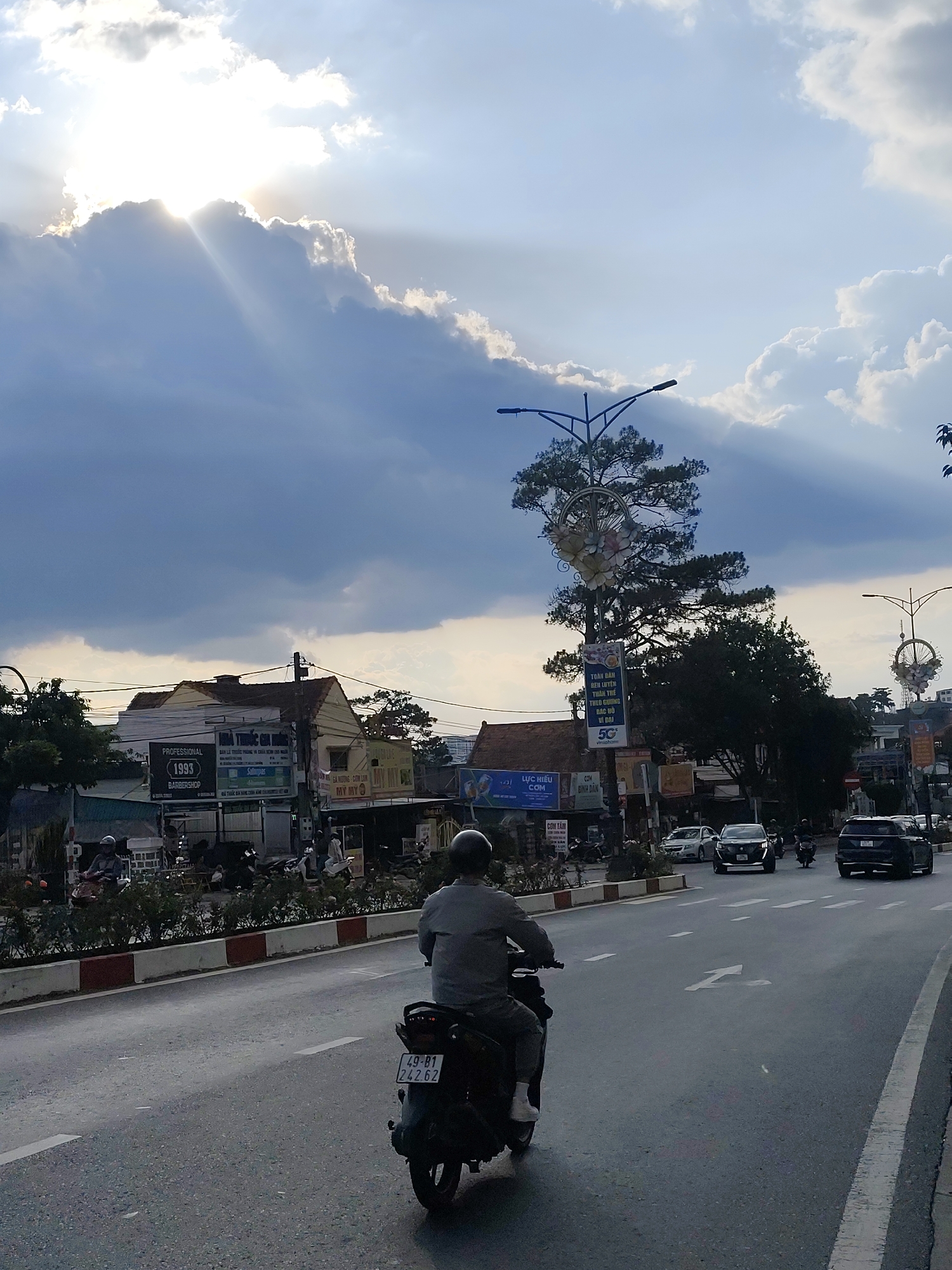 Captured in Da Lat, Vietnam, a street scene unfolds under a sky pierced by sun rays breaking through clouds. The main focus is on a lone individual riding a scooter along a well-paved road with white lane markings. The rider wears a helmet and is dressed in a gray top and dark trousers.
The surrounding urban environment includes buildings with various commercial signs written in Vietnamese, such as "PROFESSIONAL 1993 BARBERSHOP" and "LỰC HIỆU COM". A row of low bushes separates the road from the sidewalk. Several cars and other motorbikes are visible in the distance. Lampposts, adorned with floral decorations, stand along the street.