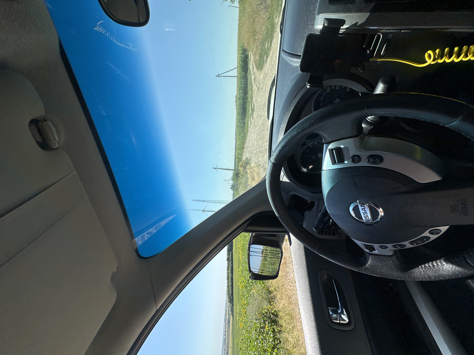 The image shows the interior of a Nissan vehicle driving on a dirt road in Korchivka, Ukraine. The driver's perspective captures the steering wheel with the Nissan logo and various dashboard controls. Outside the car, a rural landscape unfolds with a field of sunflowers on the left and a road leading through agricultural fields under a clear blue sky. Power lines and a wind turbine are visible in the distance. The sun is shining, casting shadows within the car. The overall scene depicts a typical rural drive on a sunny day.
