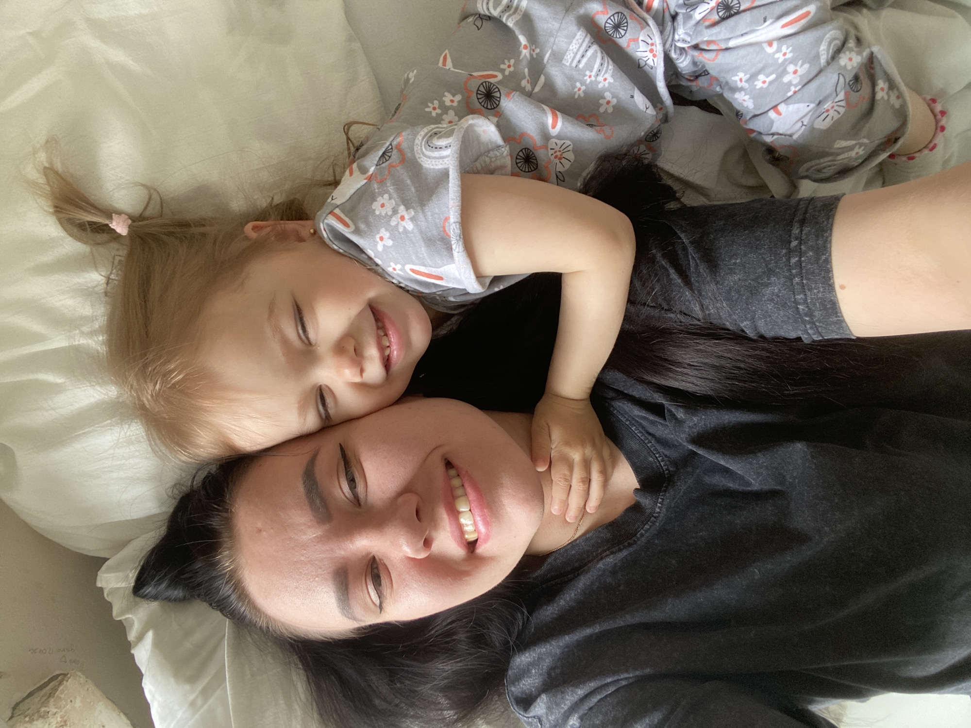 A woman and a child are lying on a white surface, possibly a bed, in an indoor setting. The woman has dark hair, dark eyebrows, and is wearing a dark gray t-shirt. She is smiling at the camera. The child, who appears to be a toddler, has blonde hair pulled back in a ponytail with a pink hair tie. The child is wearing gray pajamas with a pattern of rabbits and flowers. The child is lying on top of the woman, with one arm around the woman's neck. Both the woman and the child are smiling. The location of the image is Unknown, Unknown.