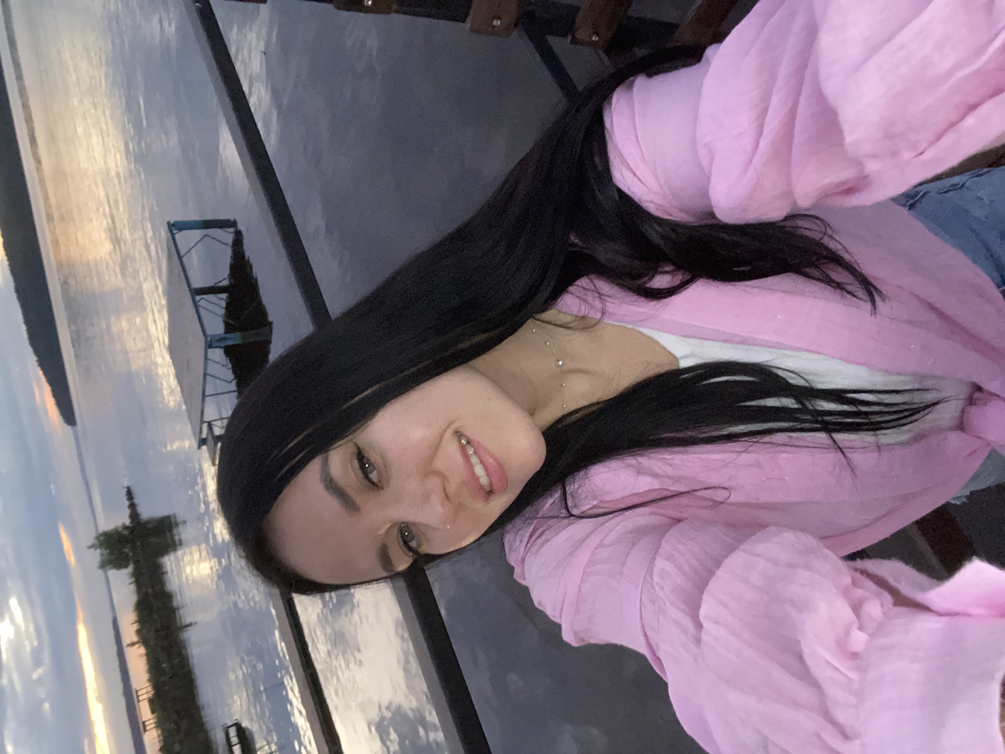 A young woman with long, dark hair takes a selfie by a body of water during dusk. She is wearing a pink top and a white undershirt, and a delicate necklace. The background shows a calm lake or river, with a distant shoreline and a cloudy sky reflecting the last light of the sun. There are structures resembling docks or platforms in the water. The overall atmosphere is serene and peaceful. The location is the embankment of the Kama River in the city of Chaikovsky, Russia.