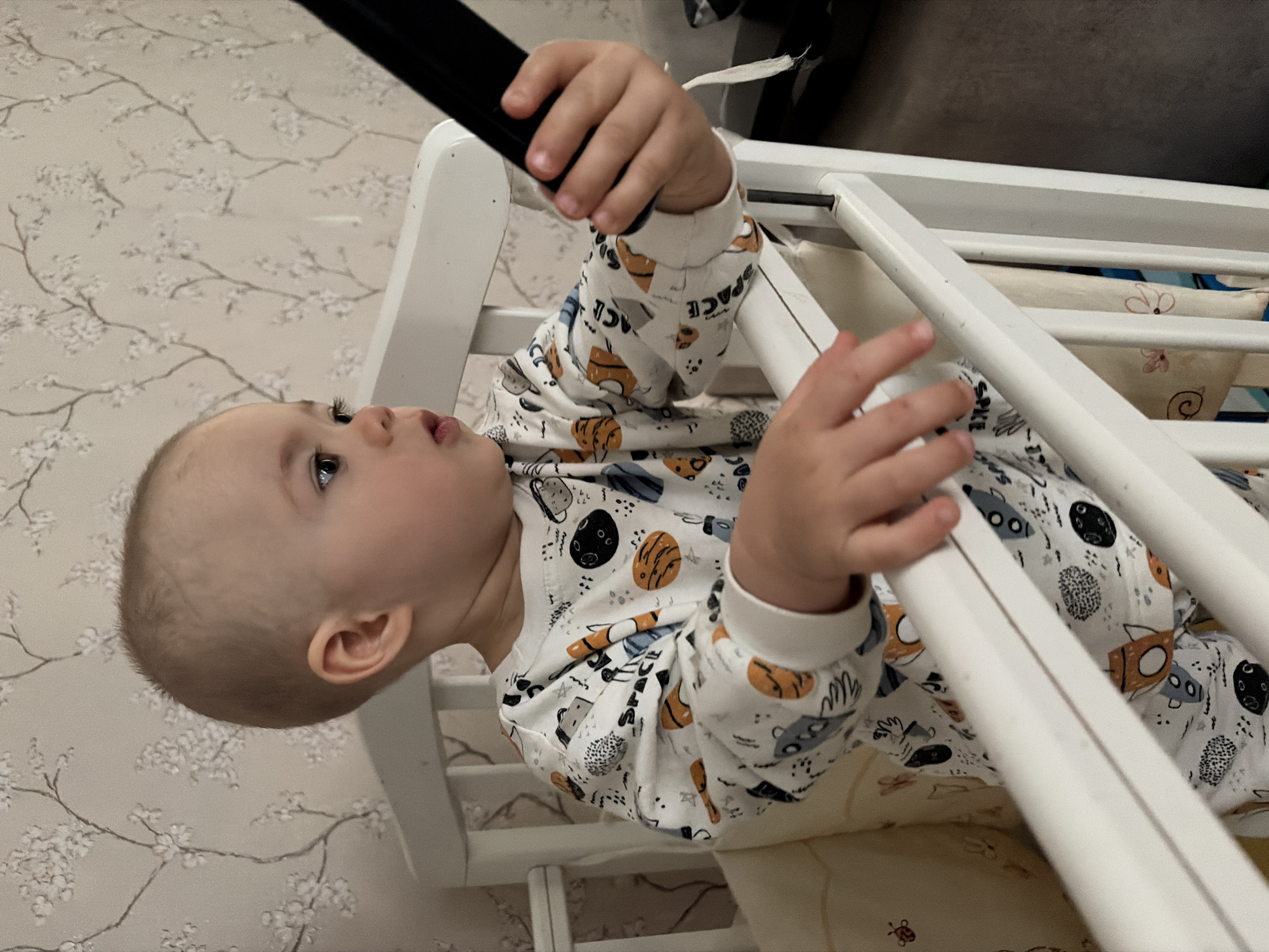A baby in a white crib with white bars and a patterned white-and-beige blanket is holding onto a black object. The baby is wearing a white long-sleeved pajama with cartoon drawings and is looking upwards. The wall behind the crib has a floral patterned wallpaper. The location appears to be indoors.