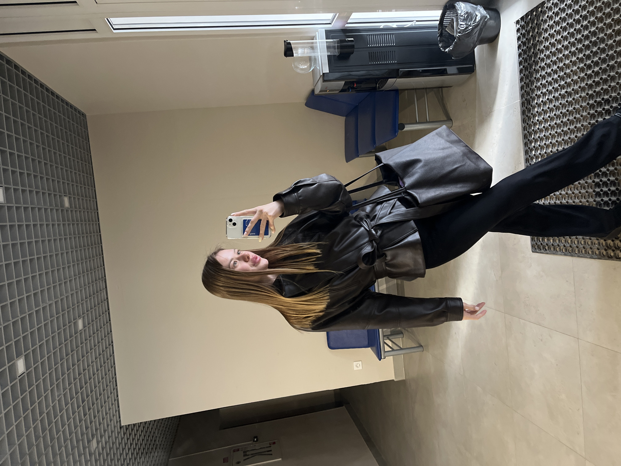 A young woman in a black leather jacket and black pants poses for a selfie in a room. She holds a smartphone in her right hand, angled towards the mirror. She has long, light brown hair with blonde highlights. A large dark-colored leather bag hangs from her shoulder. In the background, there's a tiled wall, a tan wall, blue chairs, a floor mat with a geometric pattern, and a coffee machine on a counter. The location is Chaikovsky, Russia.
