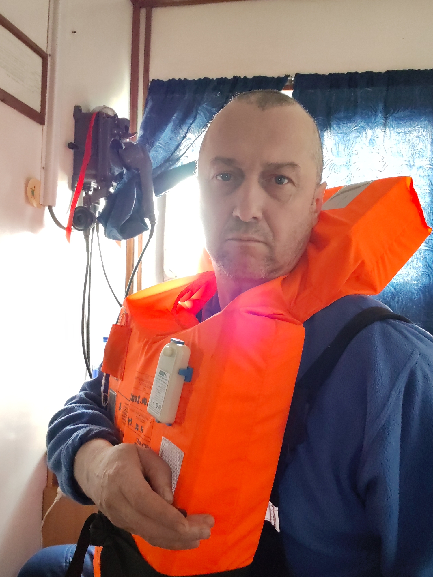 A man with a shaved head wears an orange life jacket over a blue fleece shirt. The background includes white walls, a framed picture, a vintage telephone, and blue patterned curtains. The setting appears to be the interior of a boat or ship. The location in the city of Unknown, Unknown.